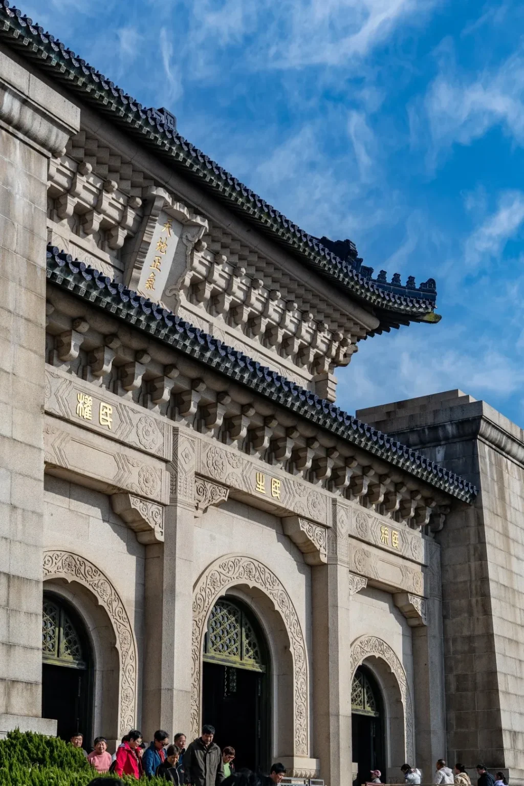 Close-up of the upper section of a large granite building with a traditional dark blue glazed tile roof. Inscriptions in gold Chinese characters are visible on the facade. Arched entrances with ornate carvings are partially visible at the bottom, with a crowd of people entering.