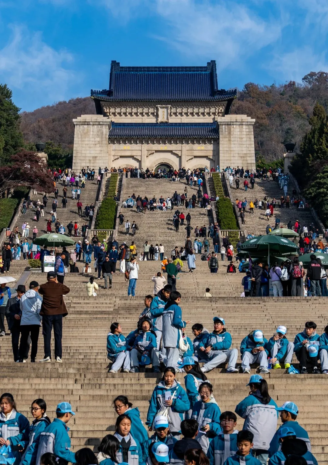 A wide view of the Sun Yat-sen Mausoleum in Nanjing, with a large stone building topped with a dark blue tiled roof at the top of a wide, long staircase. Many people, including a large group of students in matching blue and white uniforms, are visible on the steps leading up to the mausoleum. The surrounding hillside is covered in trees with autumn foliage. The sky is blue with some clouds.