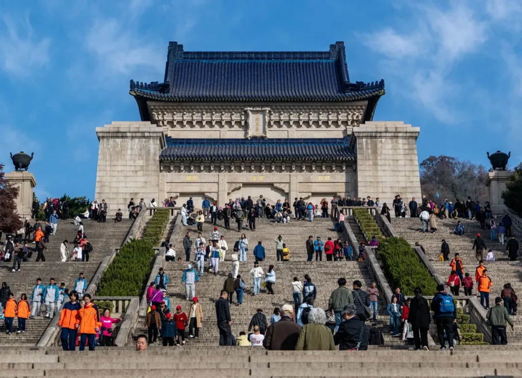 A wide, low-angle shot of a grand stone mausoleum building with a dark blue tiled roof, approached by a very wide, long, and steep set of stone stairs. Many people, dressed in casual clothing, are walking up, down, and resting on the stairs. The building features ornate architectural details and Chinese characters above a large arched entrance. The sky is bright blue with some scattered white clouds.