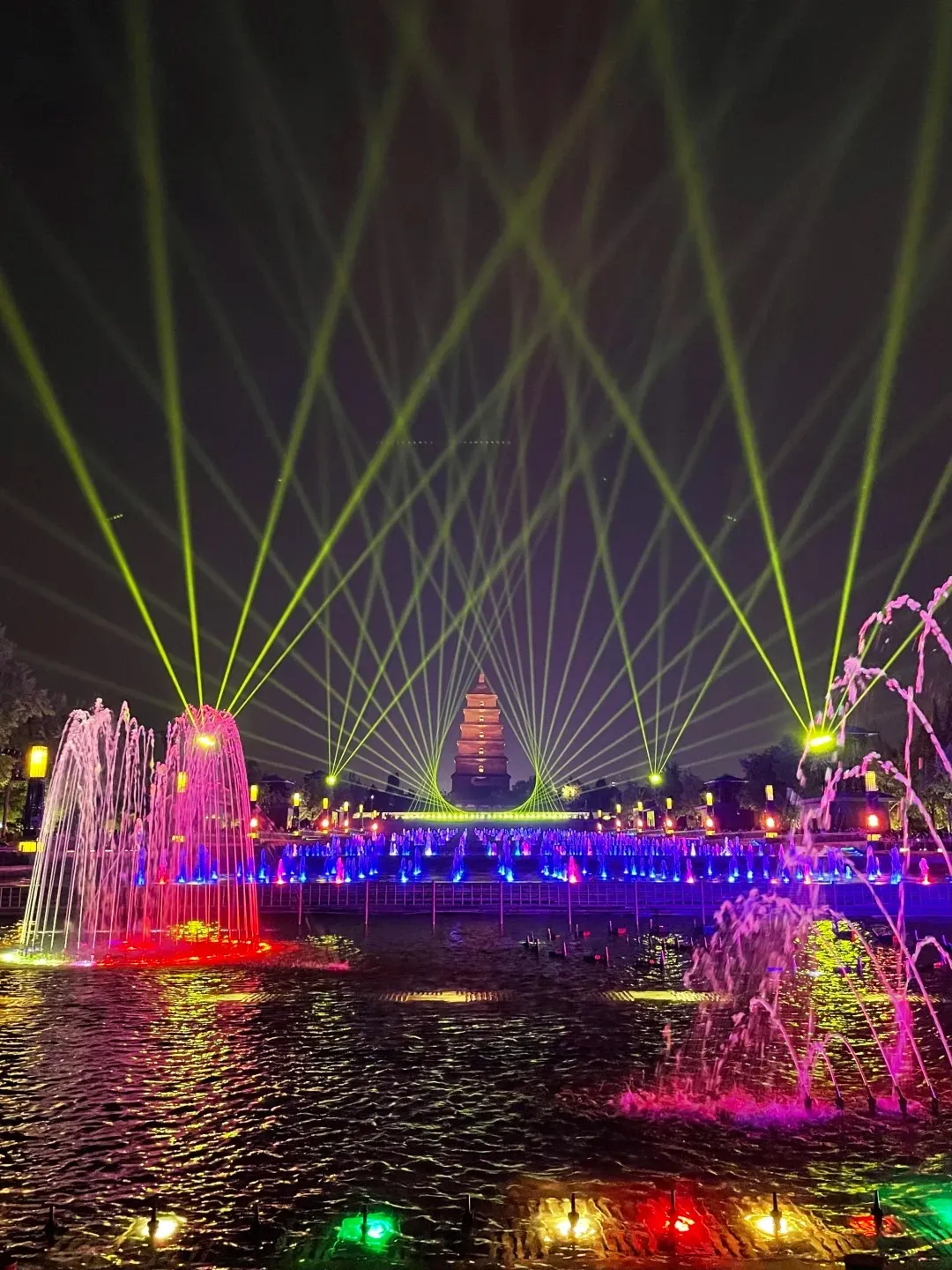 A night view of the Big Wild Goose Pagoda music fountain show in Xi'an, with brightly colored water jets in the foreground and green laser lights projecting upwards towards the pagoda.