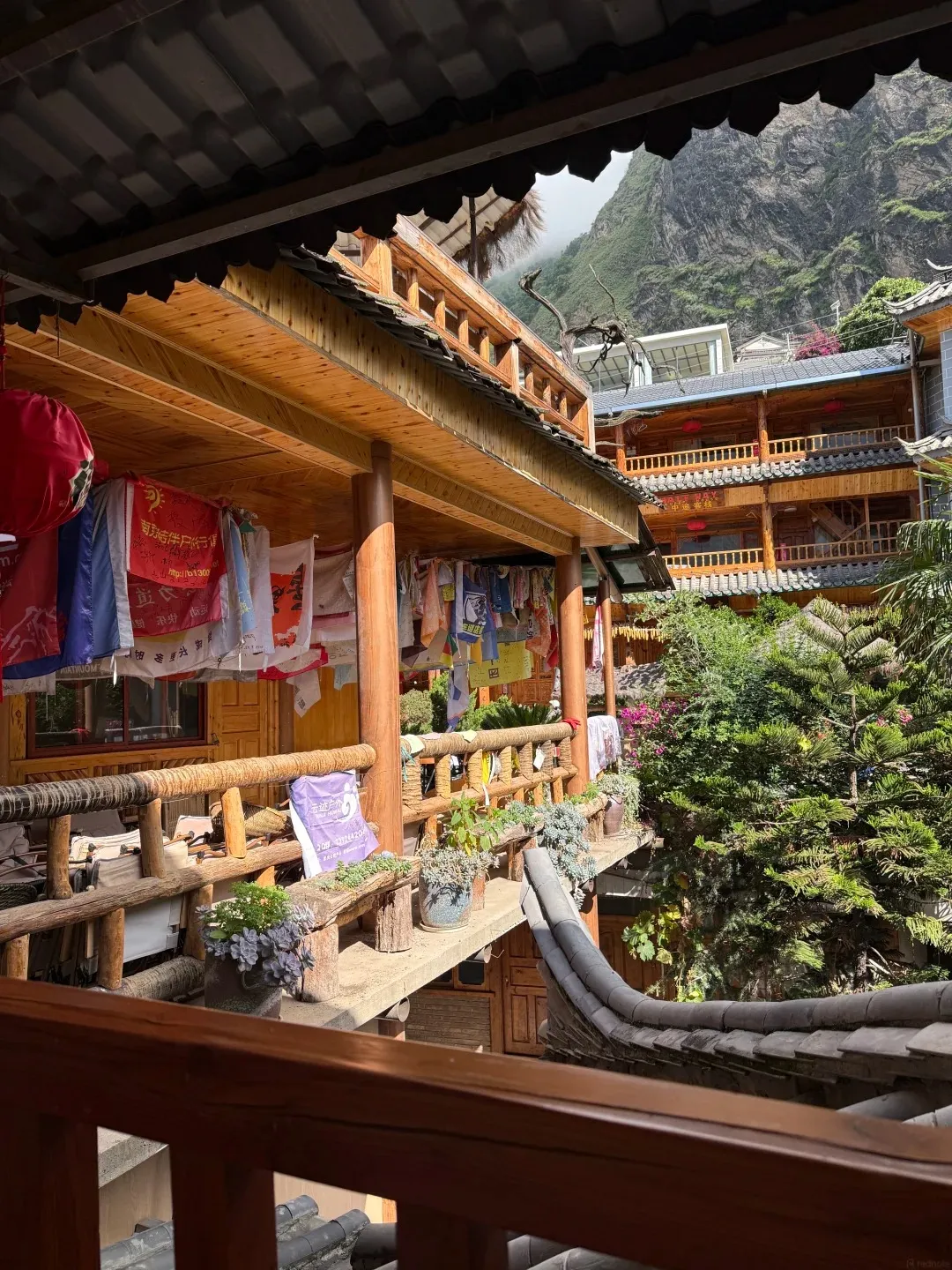 View from a wooden balcony with laundry hanging and potted plants, looking out towards a traditional multi-story wooden building with balconies, and a steep, rocky mountain covered in green vegetation in the background.
