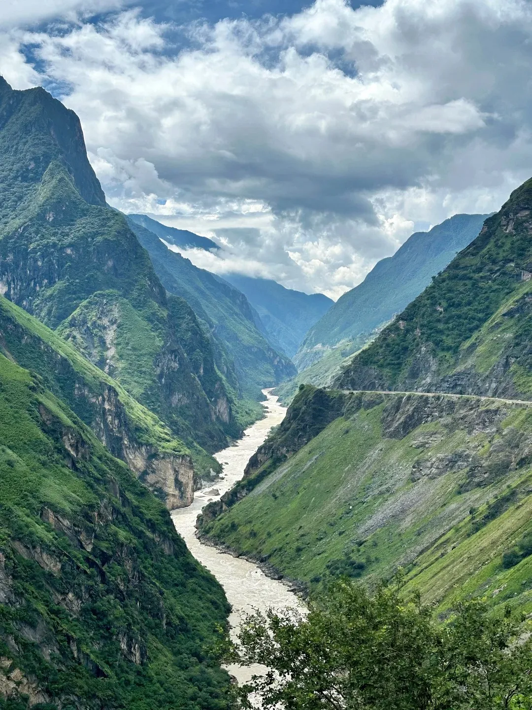 A wide, muddy river flows through a deep, narrow canyon with steep, green, vegetated mountains on either side. A dirt road with switchbacks is visible carved into the mountainside on the right.