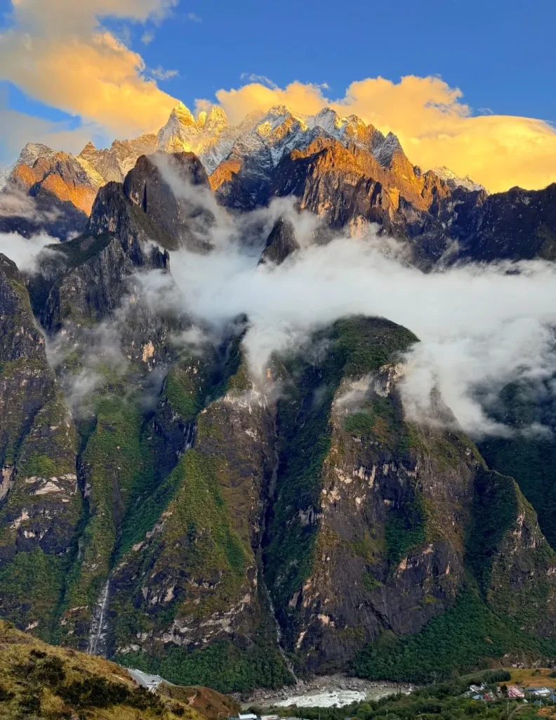 A panoramic view of Tiger Leaping Gorge at sunrise, with jagged, snow-capped mountain peaks illuminated by golden light. Clouds and mist partially obscure the steep, green-covered slopes and dark rock faces of the gorge. Far below, the Jinsha River flows through a valley with a small village visible on the right bank.