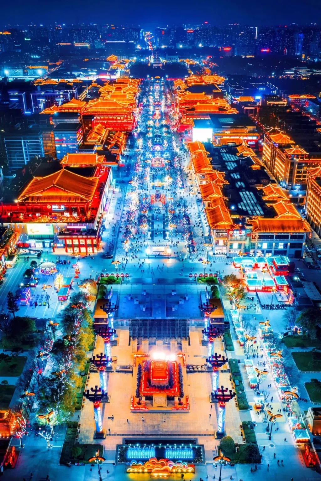 Aerial view at night of the Great Tang All Day Mall in Xi'an, with traditional Chinese architecture illuminated in orange and red. A wide pedestrian street is lined with trees decorated with colorful lights and crowded with people. In the foreground, a large plaza features decorative pillars, a central red pavilion, and a brightly lit stage.