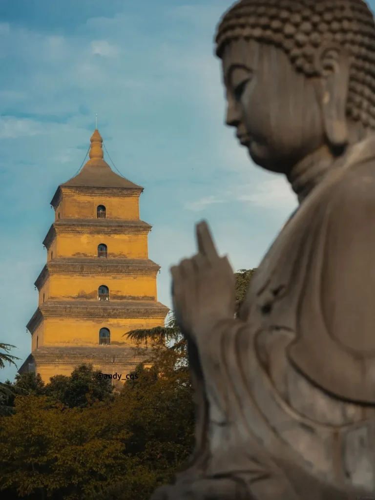A close-up, blurred foreground of a stone Buddha statue in profile, with its hand raised in a gesture. In the background, a tall, yellow, multi-tiered pagoda stands against a blue sky with scattered clouds. Green trees are visible at the base of the pagoda.