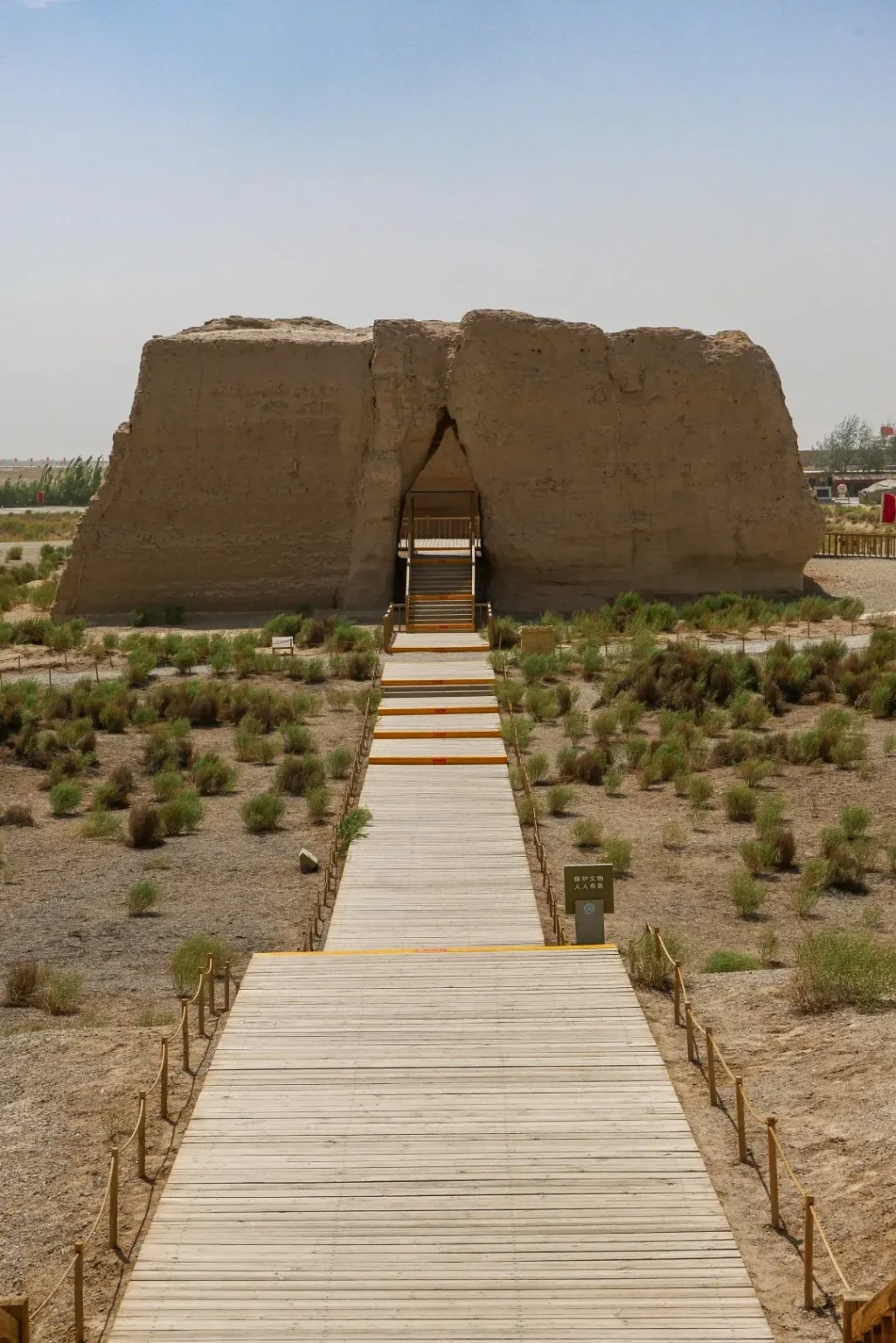 A view looking up a wooden boardwalk towards the large, trapezoidal, eroded ruins of Yumen Pass, an ancient Chinese fortress. An arched opening reveals stairs leading into the structure. The ruins are surrounded by sparse desert vegetation and a clear, pale blue sky.