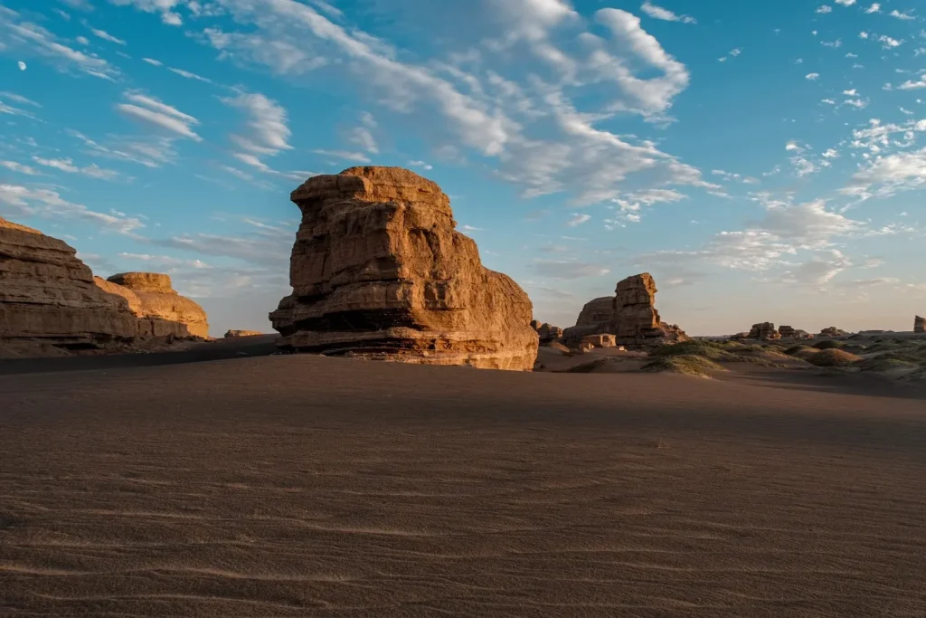 A desert landscape at sunset featuring towering, wind-eroded rock formations in warm, earthy tones. The foreground is dominated by dark sand dunes with visible ripples. The sky above is a vibrant blue with scattered wispy white clouds, and a crescent moon is visible in the upper left corner.