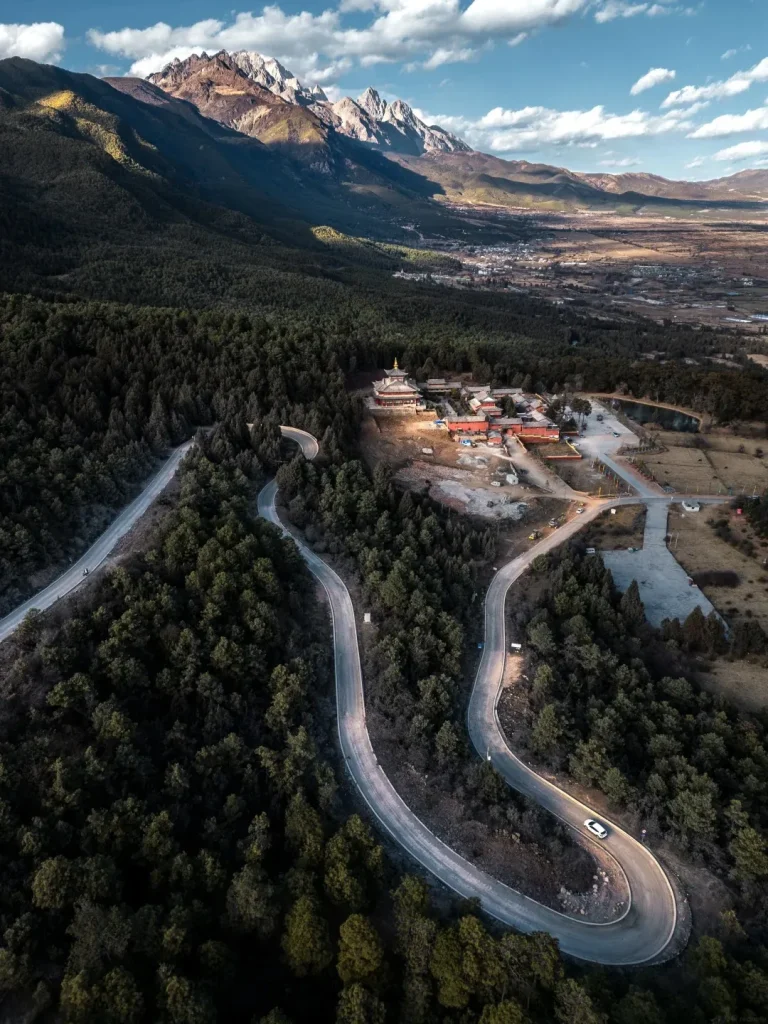 An aerial drone view of a winding road leading through a dense pine forest towards a temple complex nestled on a hillside. In the background, majestic, snow-capped mountains rise against a blue sky with scattered clouds. A small white car is visible on one of the curves of the road.