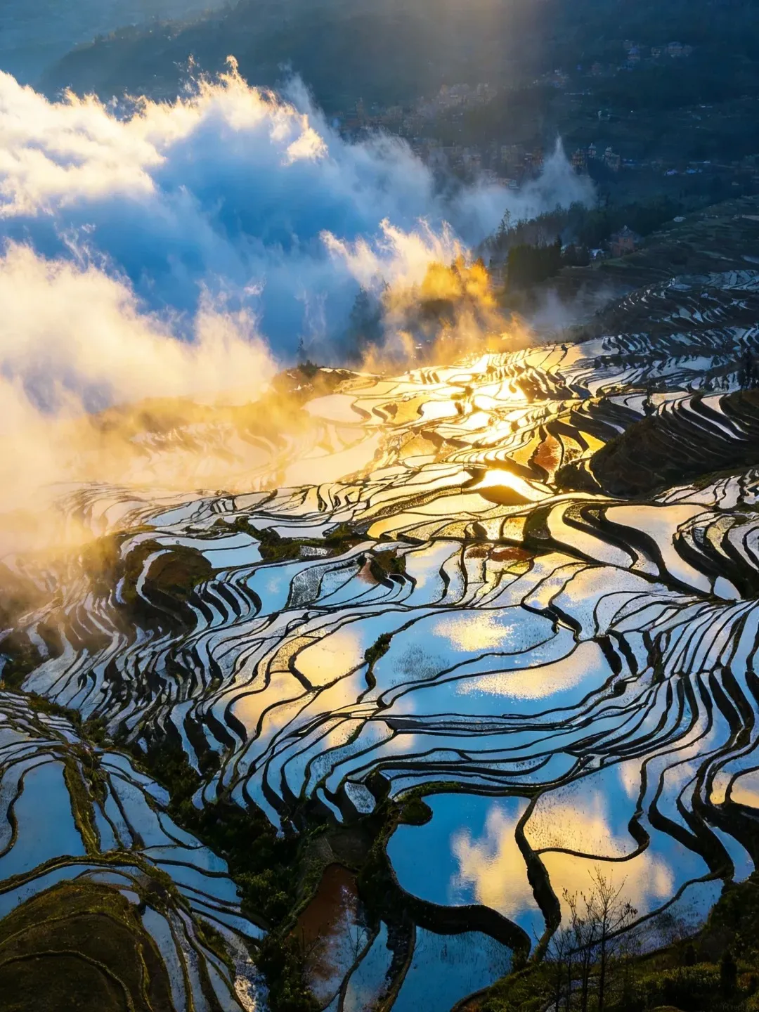 Aerial view of terraced rice paddies filled with water, reflecting the sky and golden sunlight. Clouds and mist are present in the upper part of the image, with a hint of a village on a distant hill.