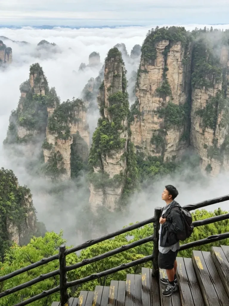A young man wearing a backpack stands on a wooden walkway with a railing, looking out at the Zhangjiajie sandstone pillars that rise through the clouds.