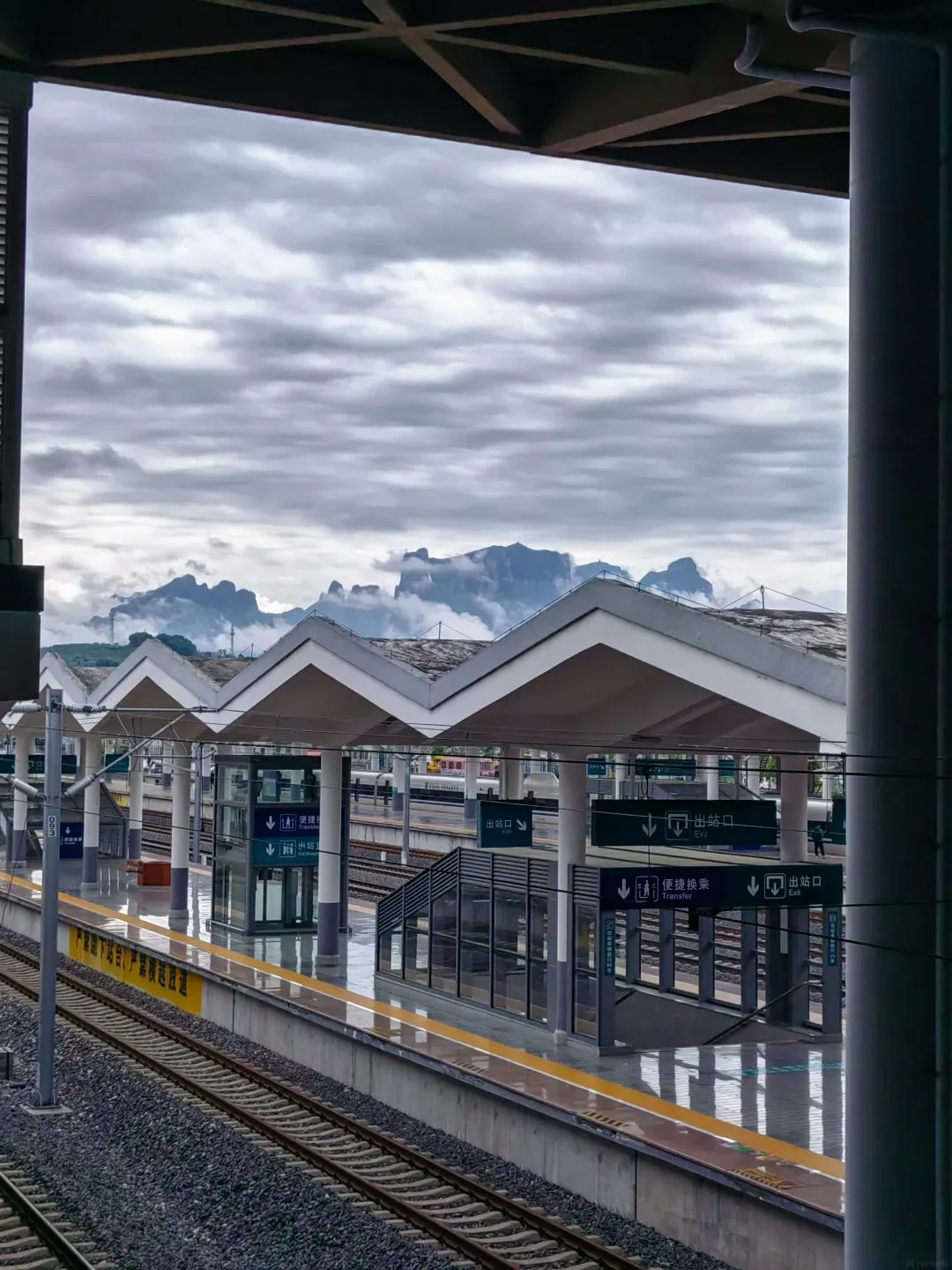 View of a modern train station platform with covered waiting areas, train tracks, and mountains in the distance under a cloudy sky. Yellow lines mark the edge of the platform.