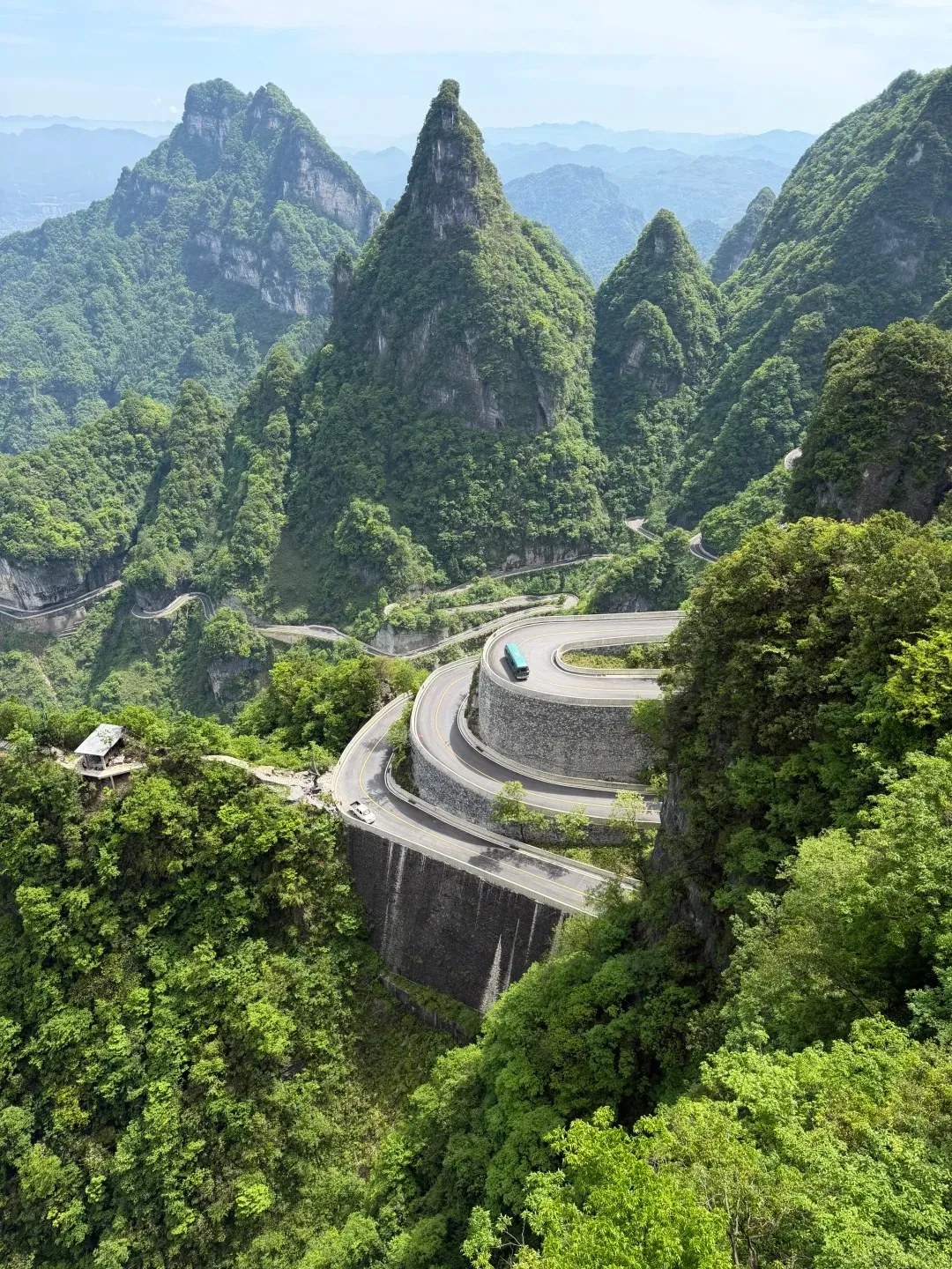 A winding, multi-layered asphalt road snakes up a steep, lush green mountain in Zhangjiajie. A teal bus and a white car are visible on the road. A small building with a red roof sits on a ledge to the left. Jagged, tree-covered peaks rise in the background under a hazy sky.
