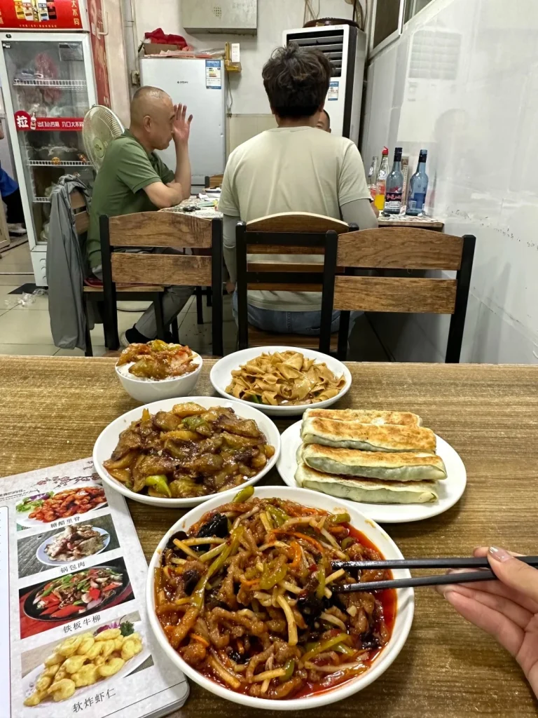 A table laden with various Chinese dishes including shredded pork with peppers, stir-fried eggplant with peppers, pan-fried dumplings, noodles, and a bowl of rice. A person's hand is using chopsticks to pick up food from the shredded pork dish. In the background, two men are seated at another table in a restaurant, with bottles of drinks on a shelf behind them.