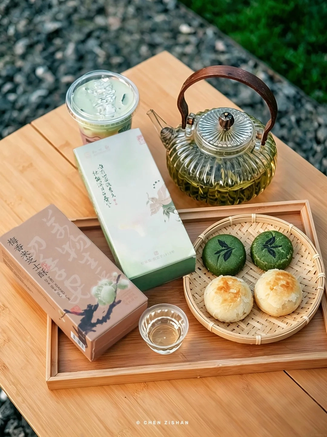 A wooden tray holds a glass teapot filled with green tea, a clear cup with a green beverage, a light brown box with Chinese calligraphy and a plum blossom illustration, a green and white box with Chinese calligraphy and a crane illustration, and a woven bamboo plate with two green pastries and two golden-brown pastries.