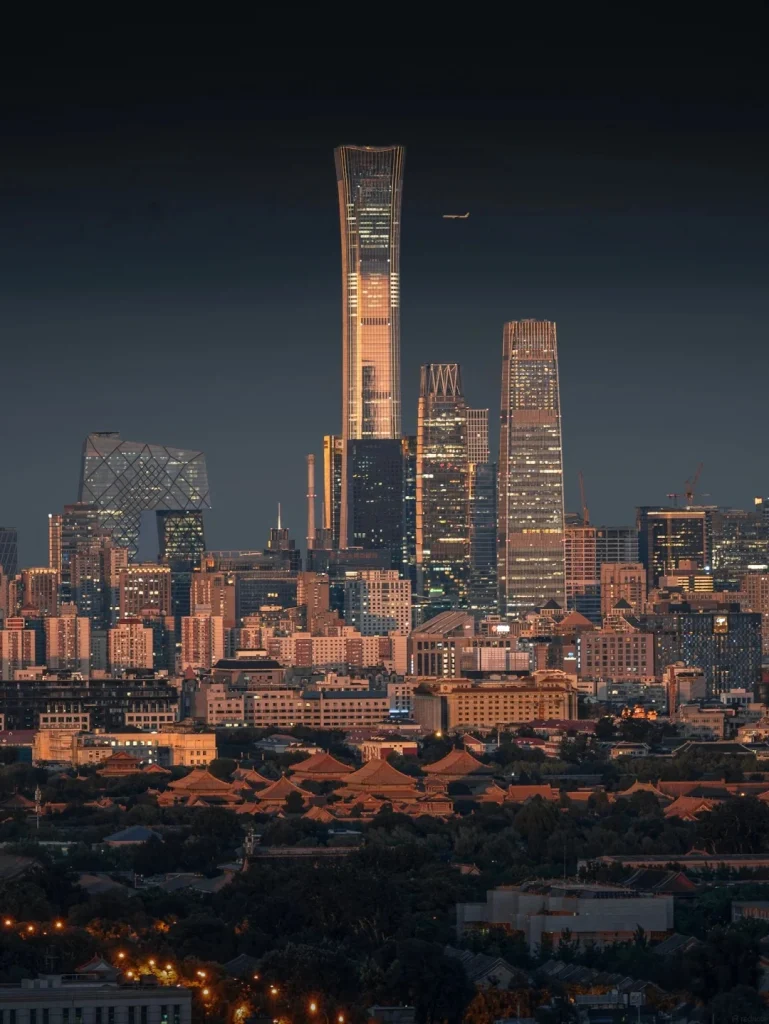 A panoramic view of a modern city skyline at dusk, dominated by towering glass skyscrapers. A distinctive curved skyscraper with illuminated windows stands prominently in the center, with an airplane visible in the dark sky above. To the left, a geometric, angular building is visible. In the foreground, traditional Chinese-style buildings with orange tiled roofs and surrounding trees create a contrast with the modern urban landscape.