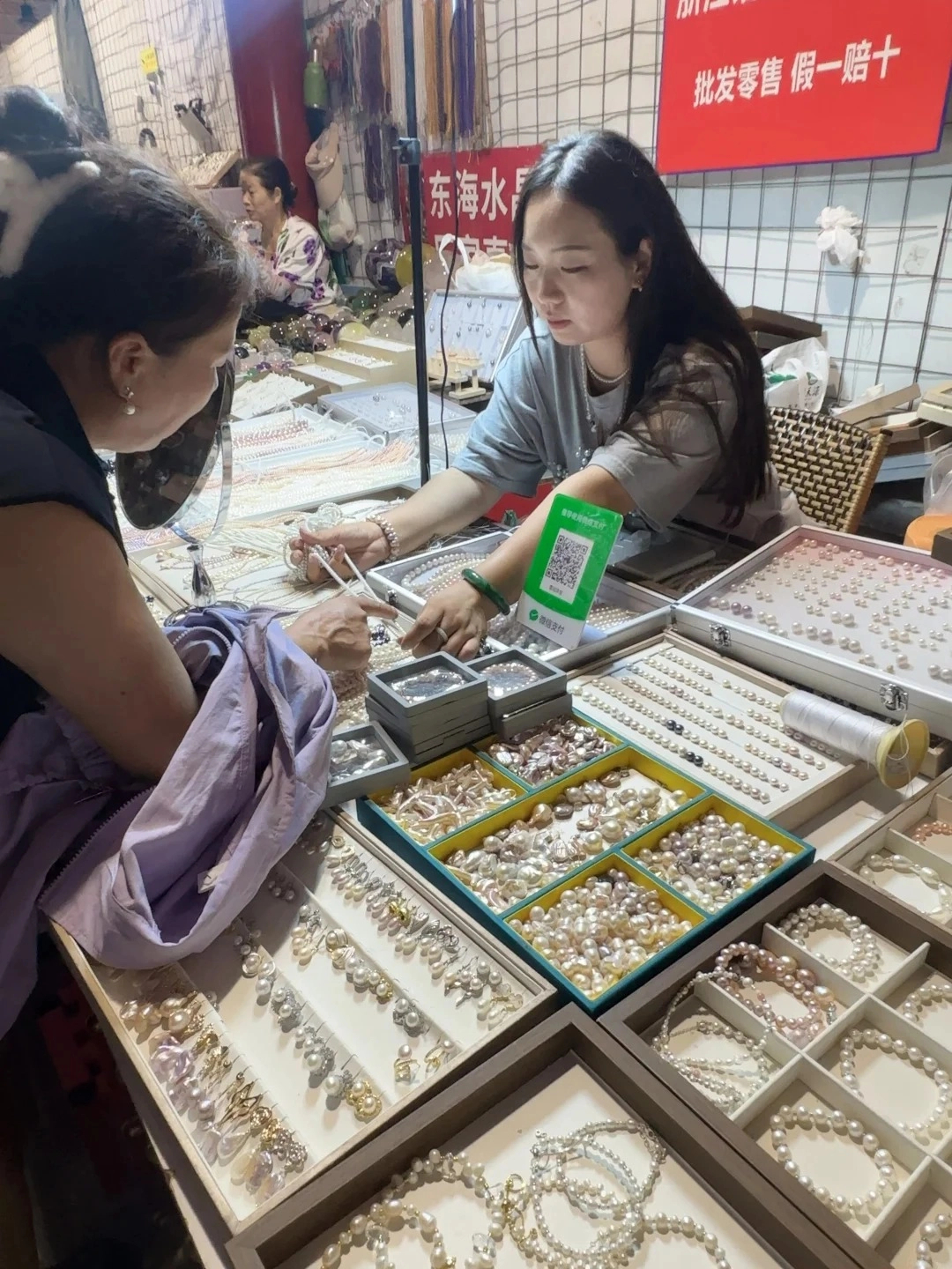 Two women are examining pearl jewelry at a market stall. The woman on the left, wearing a dark top and a light purple shawl, is looking at a display of pearl earrings and necklaces. The woman on the right, wearing a light grey t-shirt, is showing pearl necklaces to the customer and has a QR code for payment on her stall. The stall is filled with various trays of pearls, including necklaces, bracelets, and loose pearls in different colors and shapes.