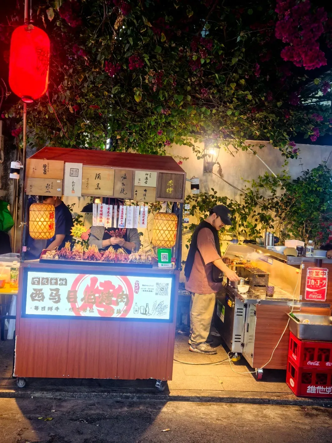 A food stall at night with a red lantern hanging above. The stall has a display of skewered food, Chinese characters on wooden boxes, and a person grilling food. A Coca-Cola crate is visible to the right.