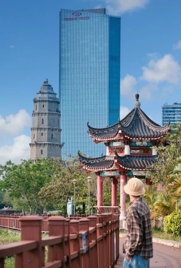 A person wearing a hat and plaid shirt stands on a walkway in front of a traditional multi-tiered pagoda with a modern blue glass skyscraper behind it. A red wooden railing runs along the walkway in the foreground.