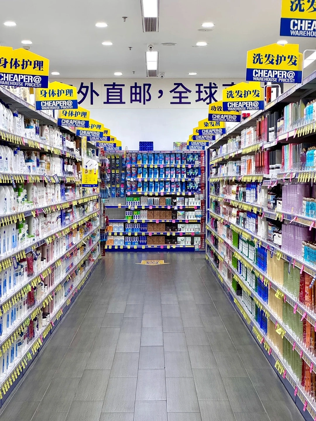 A wide shot of a brightly lit aisle in a large retail store, possibly a supermarket or discount store in China. The aisle is lined with shelves filled with health and beauty products, including shampoos, soaps, and other personal care items. Yellow signs with Chinese text and the word 'CHEAPER@' are prominently displayed hanging from the ceiling above the shelves. A large blue banner with Chinese characters runs across the top of the shelves in the background. The floor is tiled with large grey tiles, and the lighting is overhead fluorescent lights.