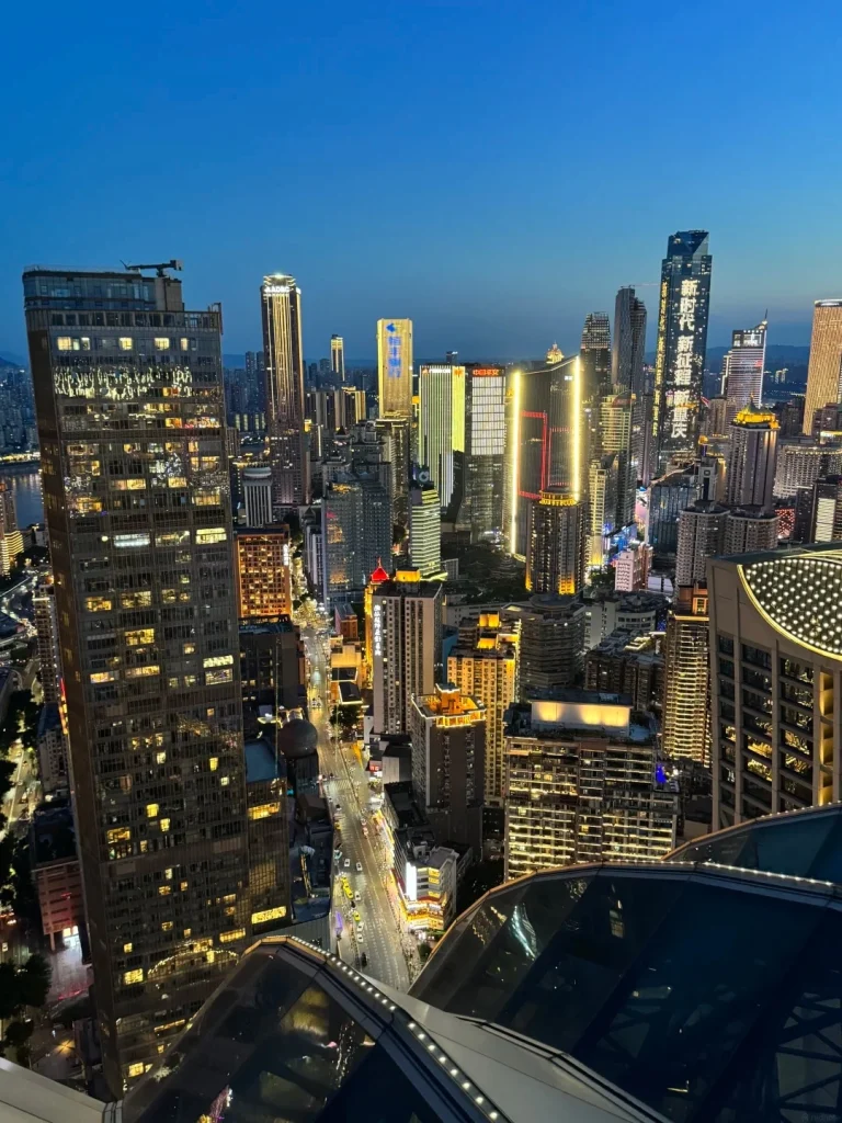 A high-angle, twilight view of a densely packed cityscape of modern skyscrapers in Chongqing, China. Many buildings are illuminated with colorful lights, including vertical bands of yellow, white, and red light on some of the taller structures. A multi-lane road with traffic and streetlights runs through the city below. The foreground features the reflective glass and metal structure of a modern building's roof or observation deck.