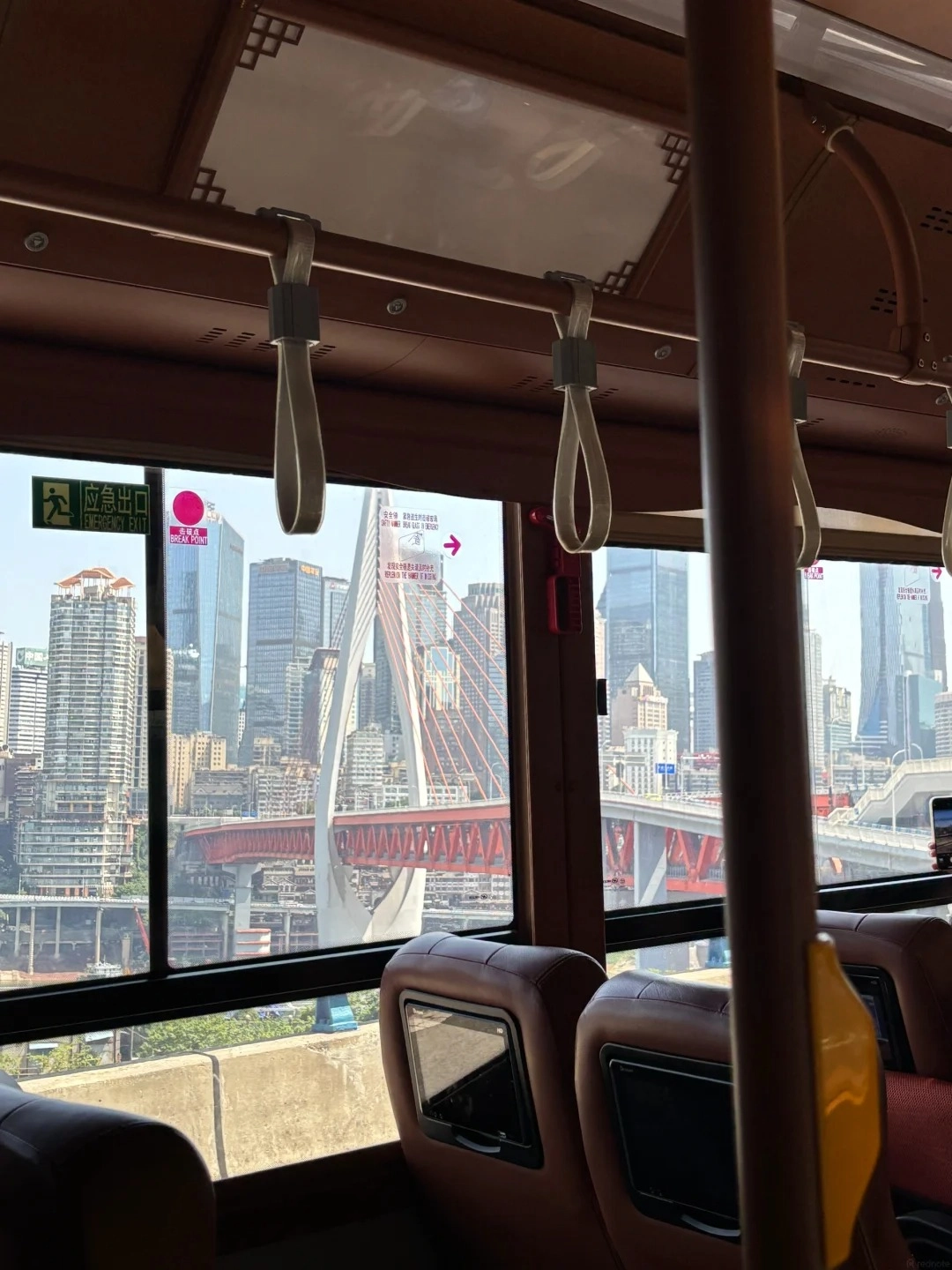 Interior view from inside a bus, looking out the window at a cityscape with tall buildings and a large red bridge. Inside the bus, there are brown leather seats and overhead handrails with white straps.