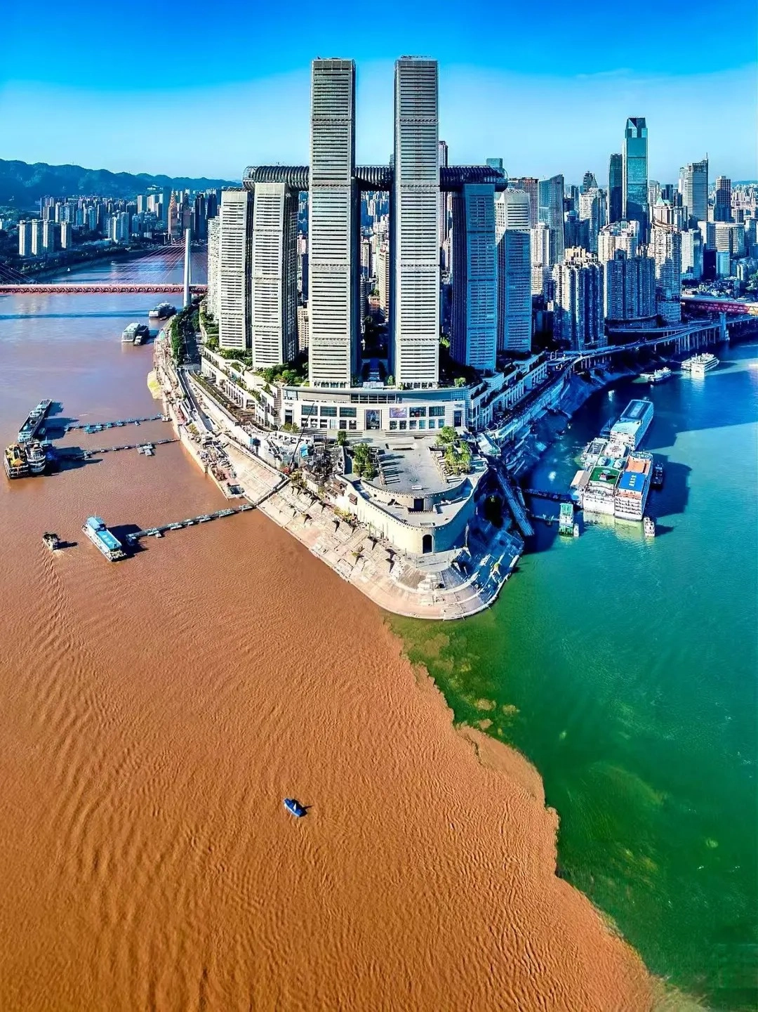 Aerial view of the confluence of two rivers, one muddy brown and the other emerald green, with the iconic Chongqing Raffles City twin skyscrapers and connecting skybridge in the foreground, and a cityscape with bridges and buildings in the background under a clear blue sky.