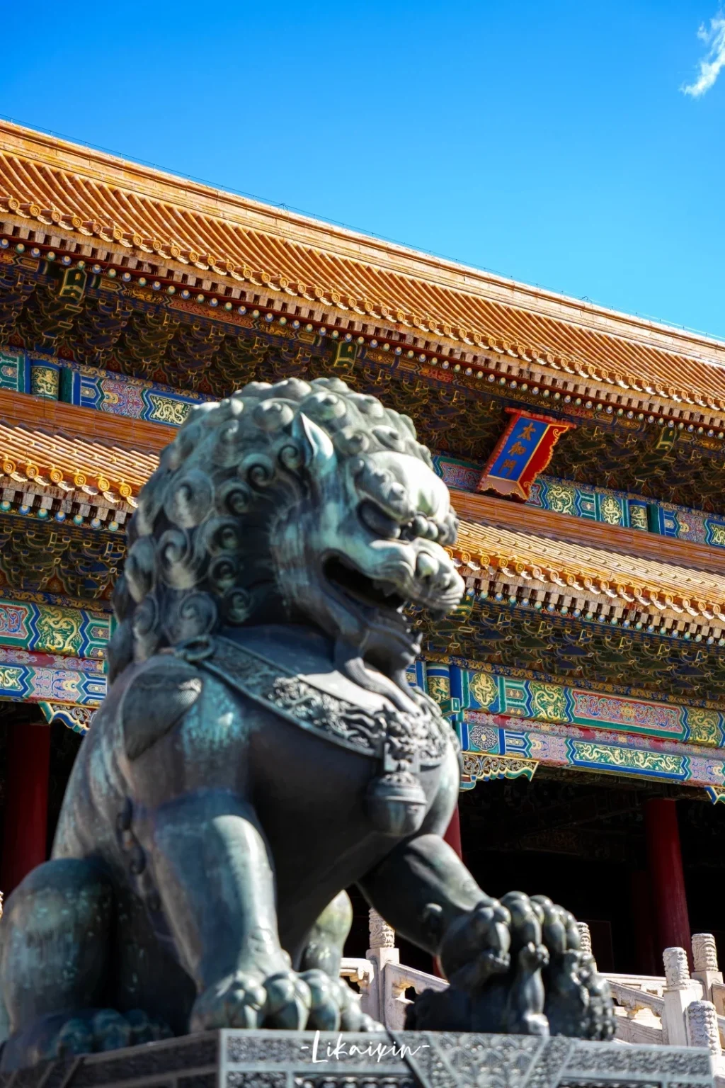 A close-up, low-angle view of a weathered bronze lion statue in front of the ornate, yellow-tiled roof and colorful painted eaves of the Forbidden City in Beijing, China. The sky above is a clear, bright blue.