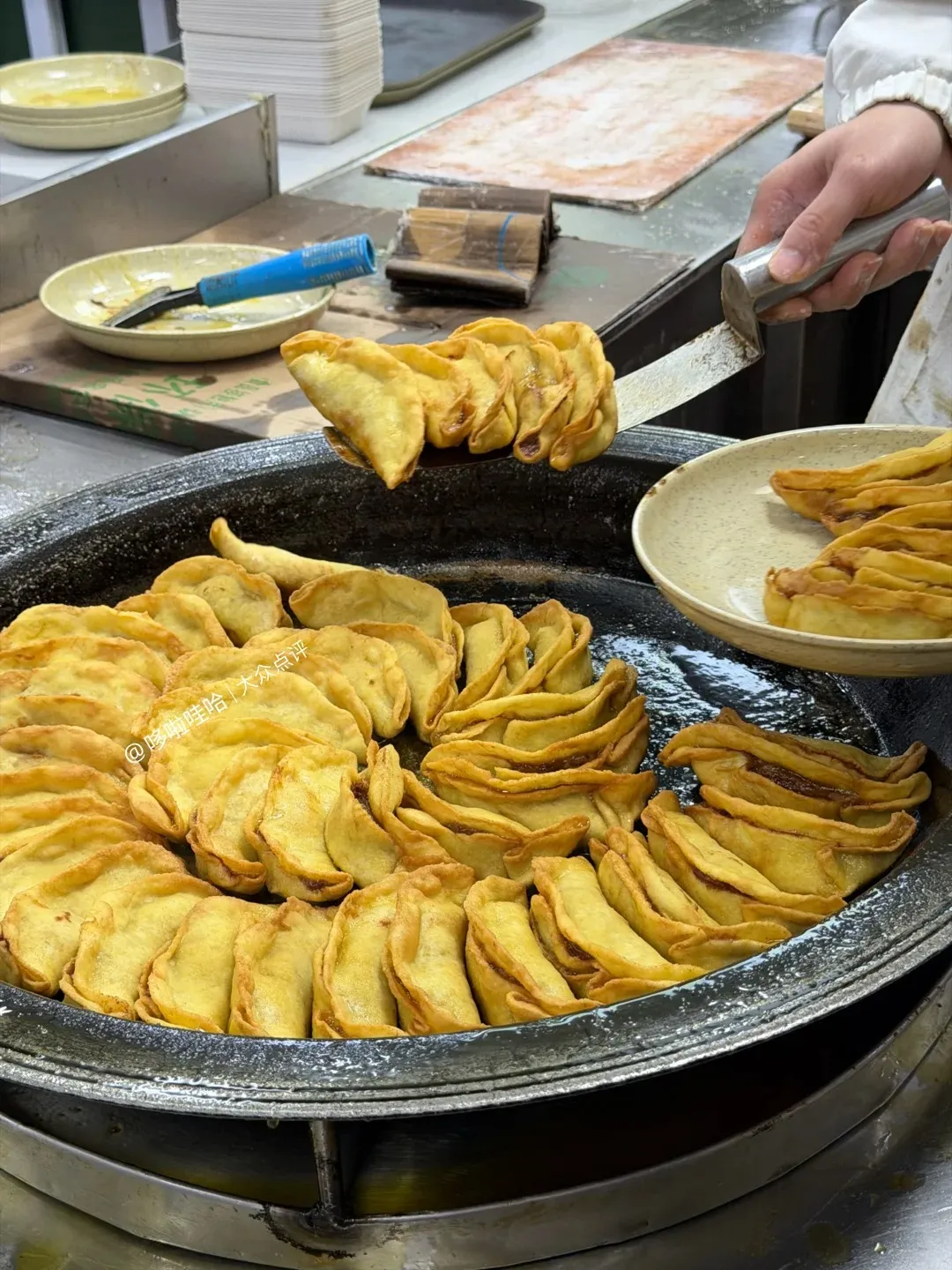 A close-up shot of golden-brown, pan-fried dumplings arranged in a large, round, dark-colored pan. A spatula is lifting a portion of the dumplings, showing their pleated edges and filling. Another plate of fried dumplings is visible to the right.
