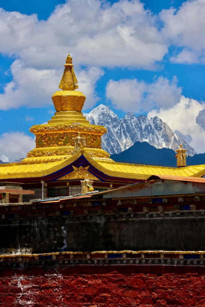 A large golden stupa rises above the yellow, blue, and red tiled roof of Lhagong Monastery, with snow-capped mountains in the background under a bright blue sky with white clouds.