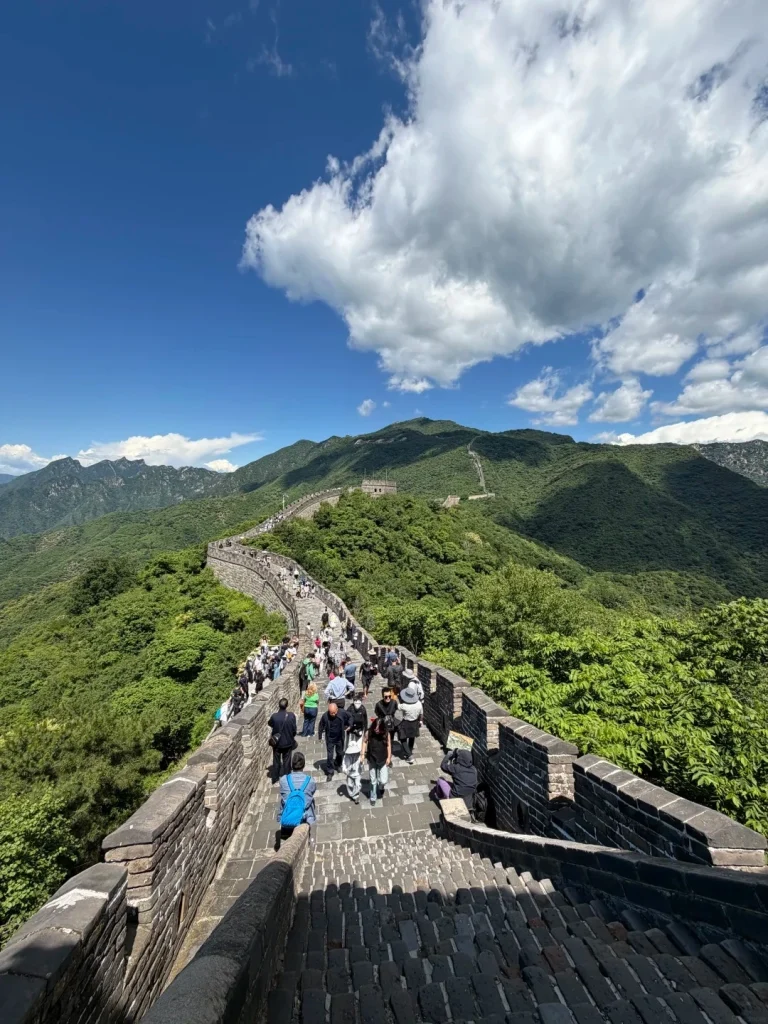 A perspective shot looking down a section of the Great Wall of China. A long line of tourists walk along the wall, which is flanked by lush green mountains on either side under a bright blue sky with scattered white clouds. The wall itself is made of stone bricks and has crenellated battlements.