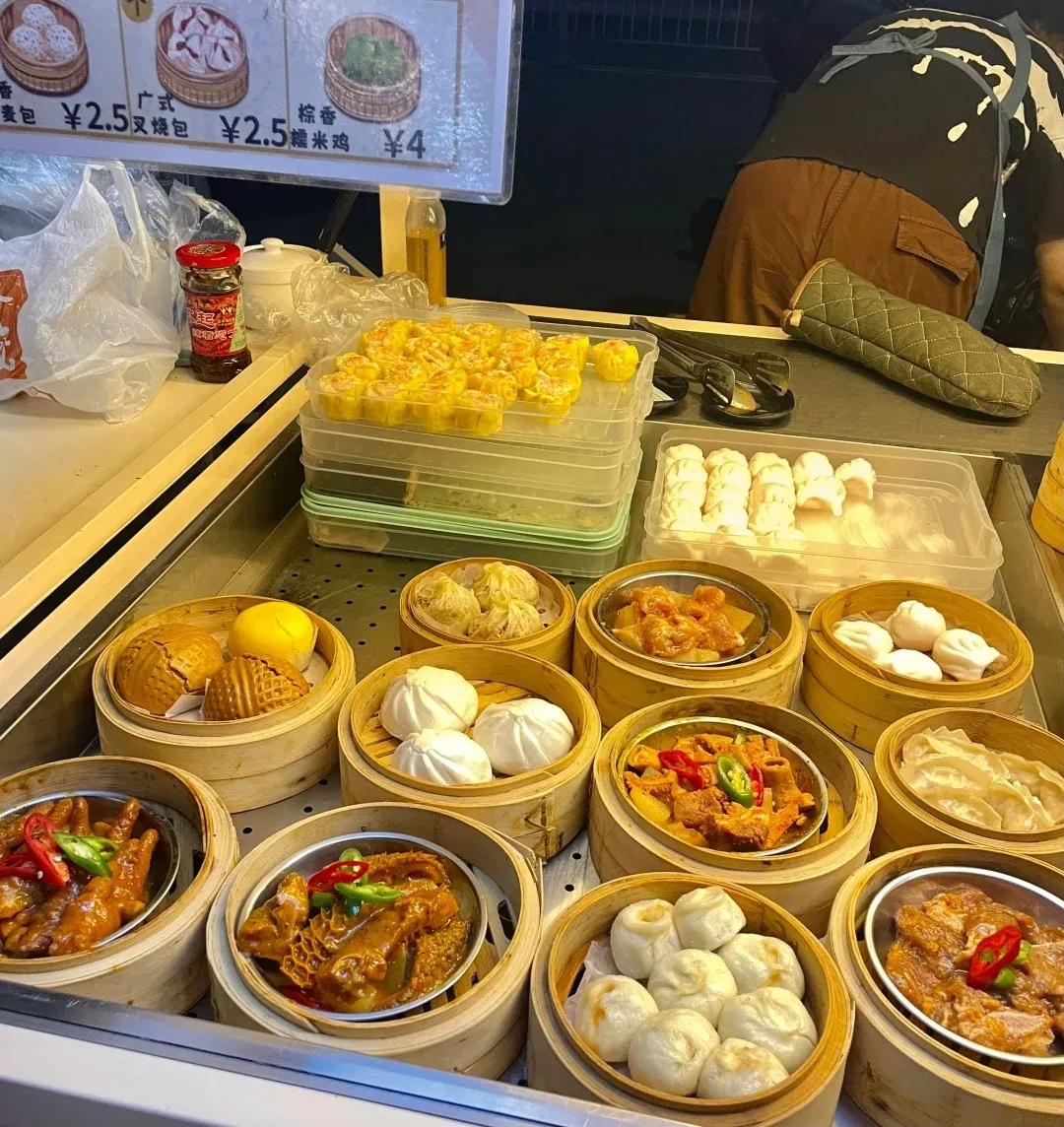 A close-up view of various Cantonese dim sum and steamed dishes displayed in bamboo steamers on a metal counter. In the foreground, there are several bamboo steamers filled with different food items. The leftmost steamer contains braised chicken feet with chili and sauce. Next to it is a steamer with tripe in a rich brown sauce. Moving right, there are steamers with white steamed buns, small steamed dumplings (shumai), and a dish of diced meat with vegetables. In the upper part of the image, there are trays filled with small, yellow steamed dumplings or sui mai. A small bottle of red condiment is visible on the left. In the background, a person wearing an apron is partially visible.