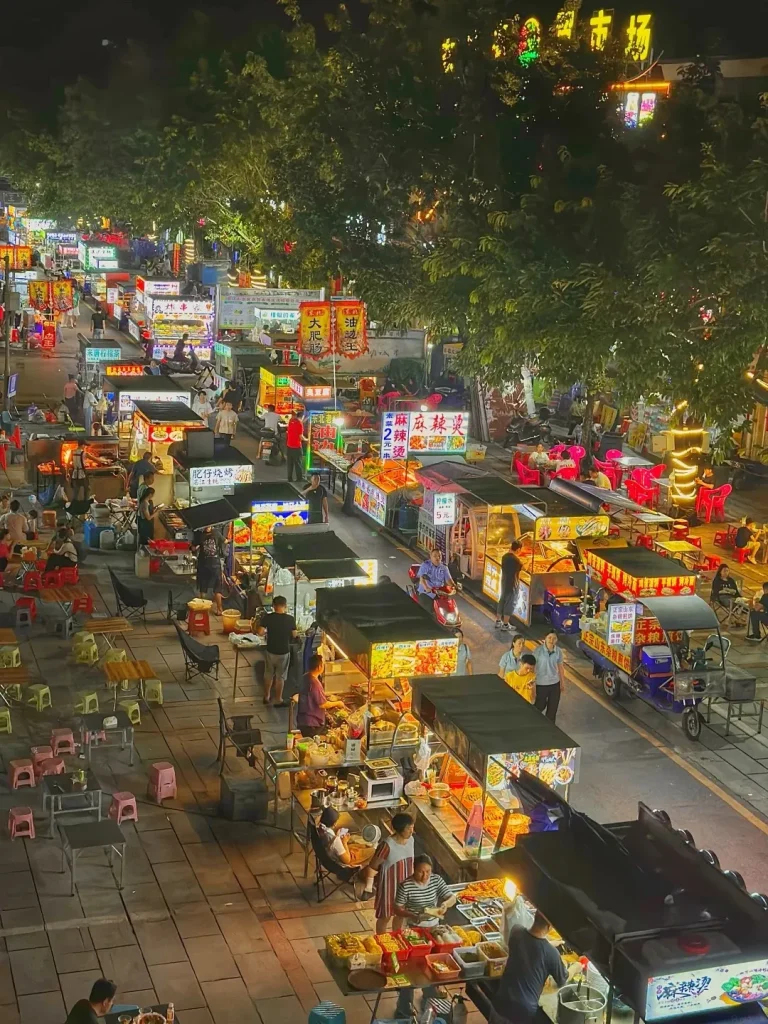 An aerial view of a busy Guangzhou night market at night, with numerous illuminated street food stalls lining a street. People are seen walking, eating, and interacting at the stalls, with trees and lights overhead. Some stalls have colorful signs and displays of food, while others are equipped with cooking equipment. Small tables and chairs are set up for diners. Several motorized carts, including a blue three-wheeled vendor vehicle, are also present on the street.
