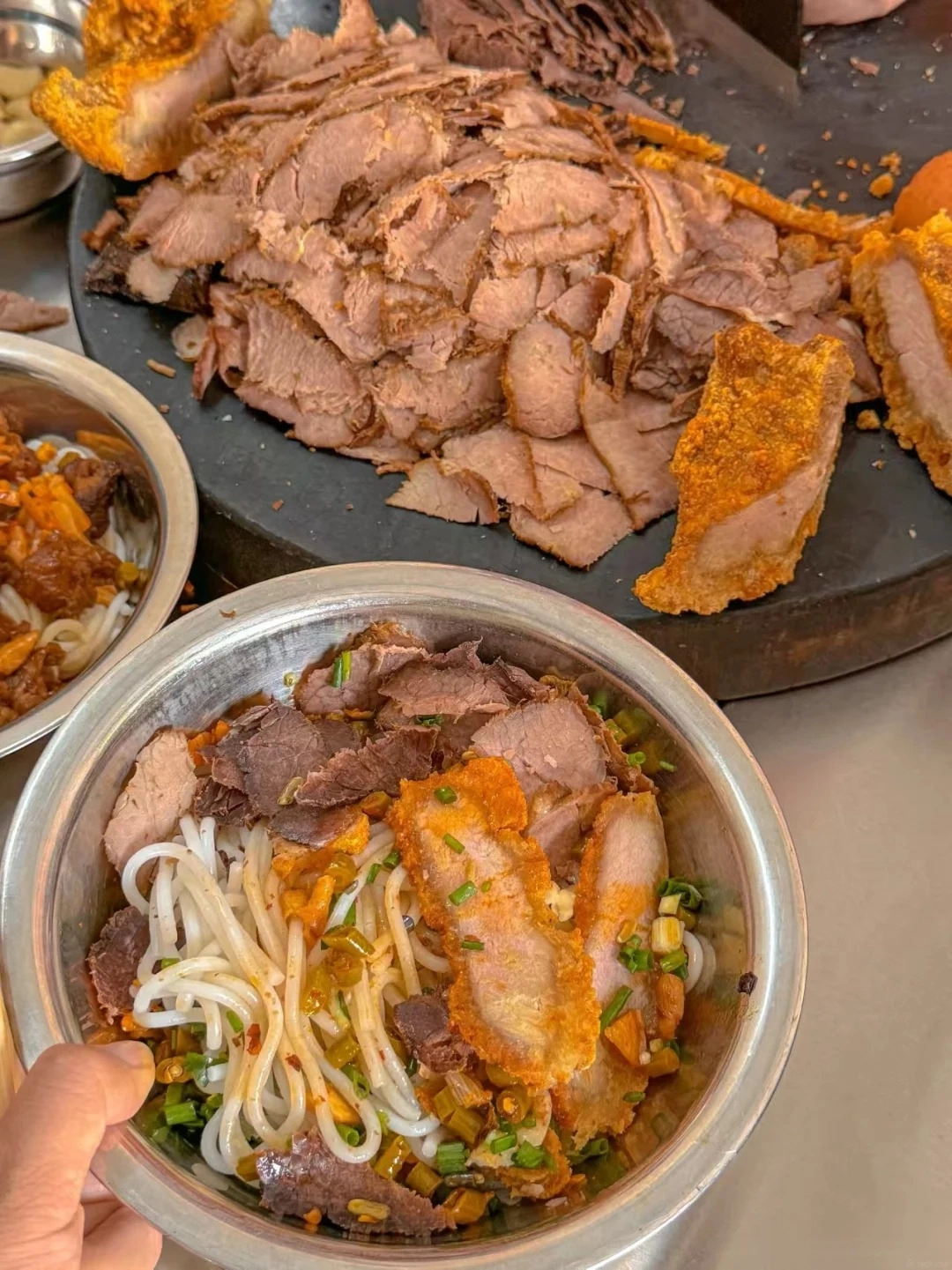 A close-up of a metal bowl filled with white rice noodles, topped with sliced roasted beef, pieces of crispy pork belly, chopped green onions, and a savory sauce with small green pepper pieces.