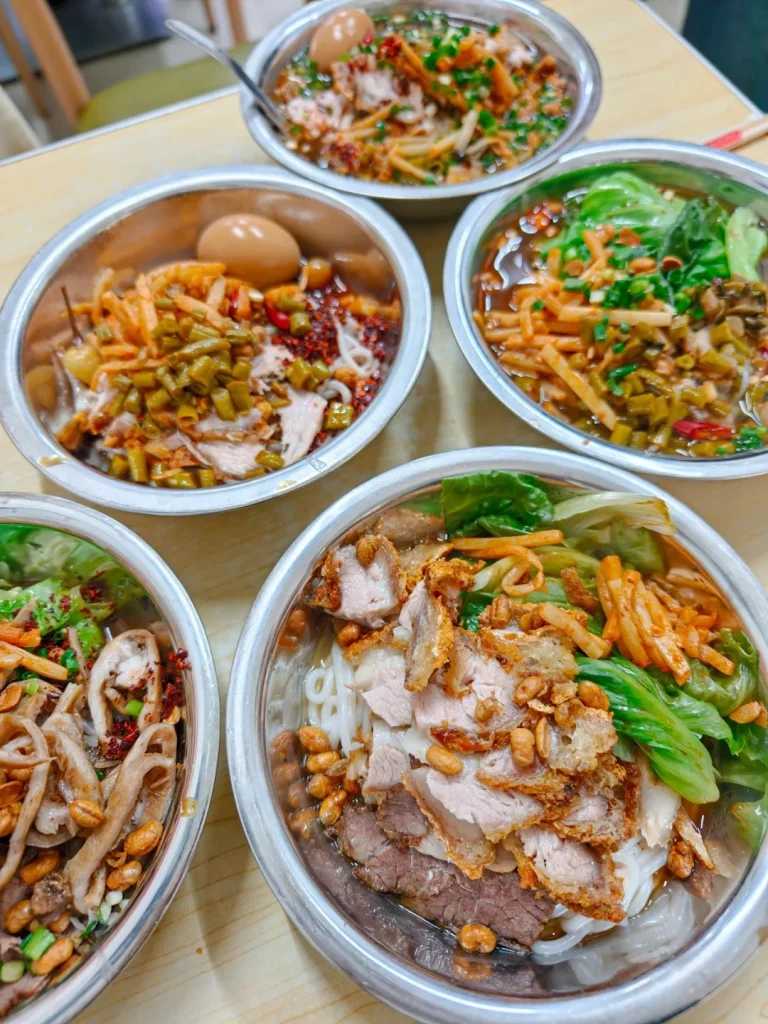 A close-up overhead view of four bowls of Guilin rice noodles on a wooden table. The bowl in the foreground contains thick noodles topped with slices of dark beef, crispy pork belly, pickled vegetables, peanuts, and lettuce. The other bowls contain noodles with various toppings including eggs, pickled vegetables, meat, and herbs, with some bowls showing broth.