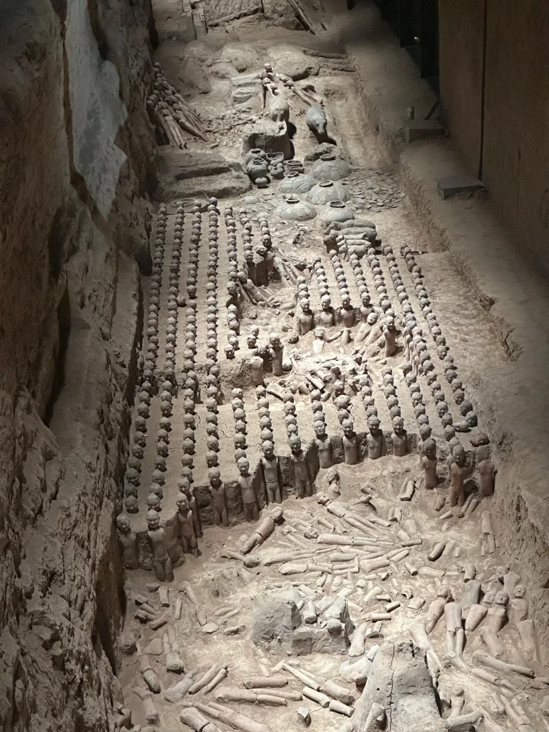 An elevated view of an archaeological excavation pit at the Han Yang Ling Museum, revealing rows of miniature terracotta figures and scattered fragments of pottery and other artifacts. A walkway with a glass floor allows visitors to observe the site from above.