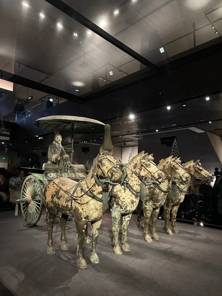 A life-sized bronze chariot pulled by four horses, with a terracotta figure of an attendant seated on the chariot under a large umbrella. The chariot and horses are displayed on a dark gray surface with theatrical lighting, and a glimpse of museum visitors can be seen in the background.
