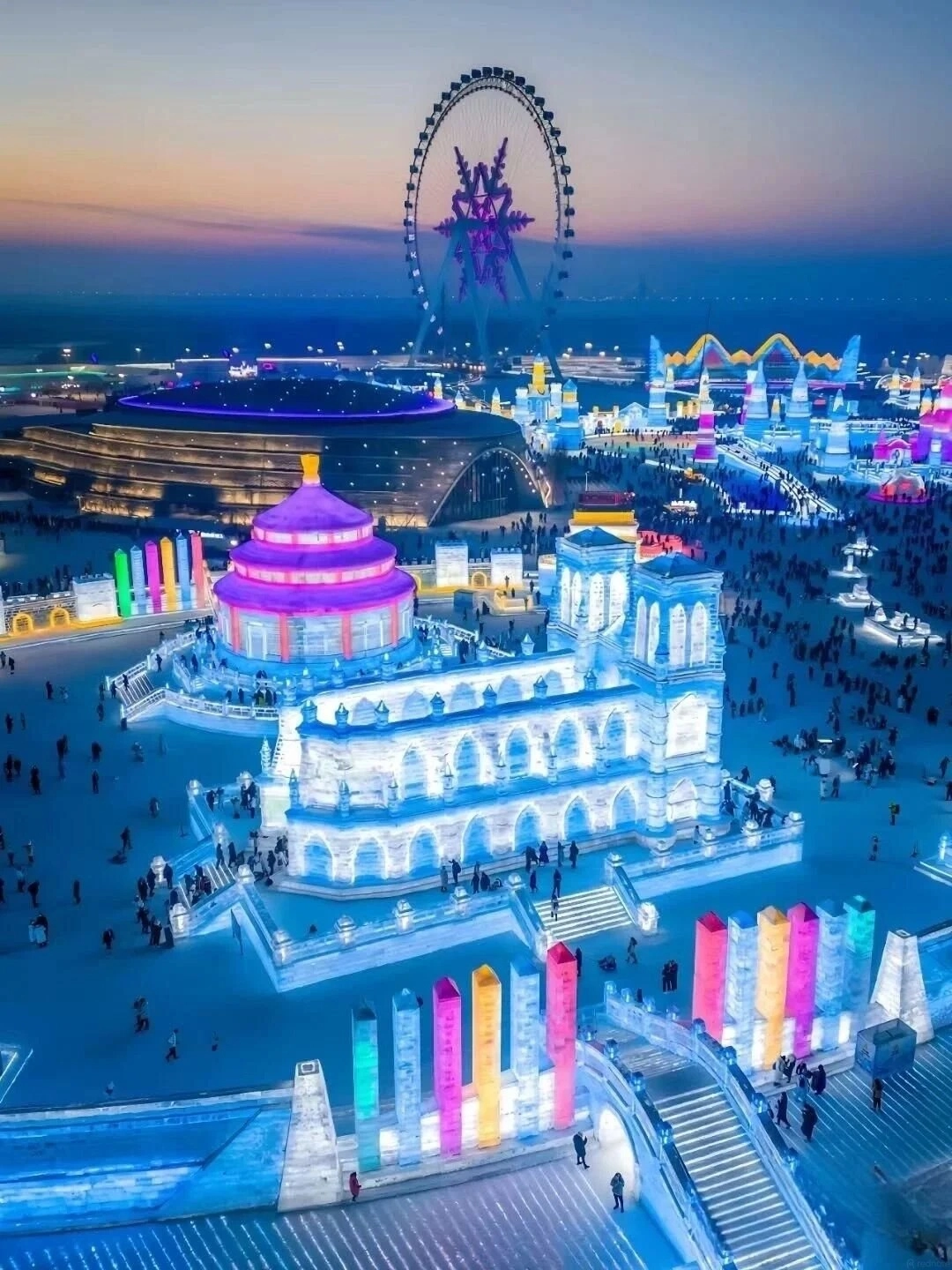 An aerial view of a nighttime winter festival with brightly illuminated ice sculptures and buildings in various colors, including a large ice castle structure with turrets and arches, a domed building with purple lighting, and numerous smaller ice structures. A large Ferris wheel with snowflake-shaped lights is visible in the background, and a crowd of people are walking around the illuminated park. The sky above is a gradient of dark blue and orange.
