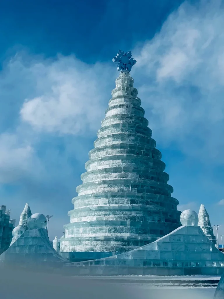 A large, tiered ice sculpture shaped like a Christmas tree with a snowflake on top, set against a bright blue sky with white clouds. Smaller ice sculptures are visible in the foreground and to the sides.