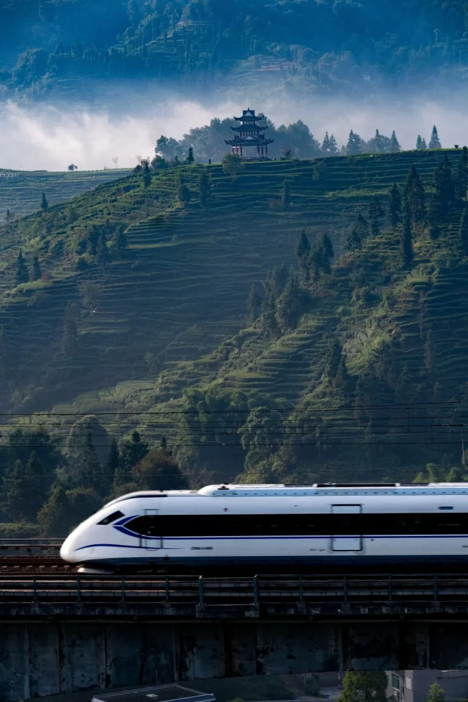 A white and blue high-speed train travels on tracks elevated on a bridge, passing in front of a steep, mist-covered hillside covered in lush green tea terraces. A traditional multi-tiered Chinese pagoda sits atop the hill in the distance.