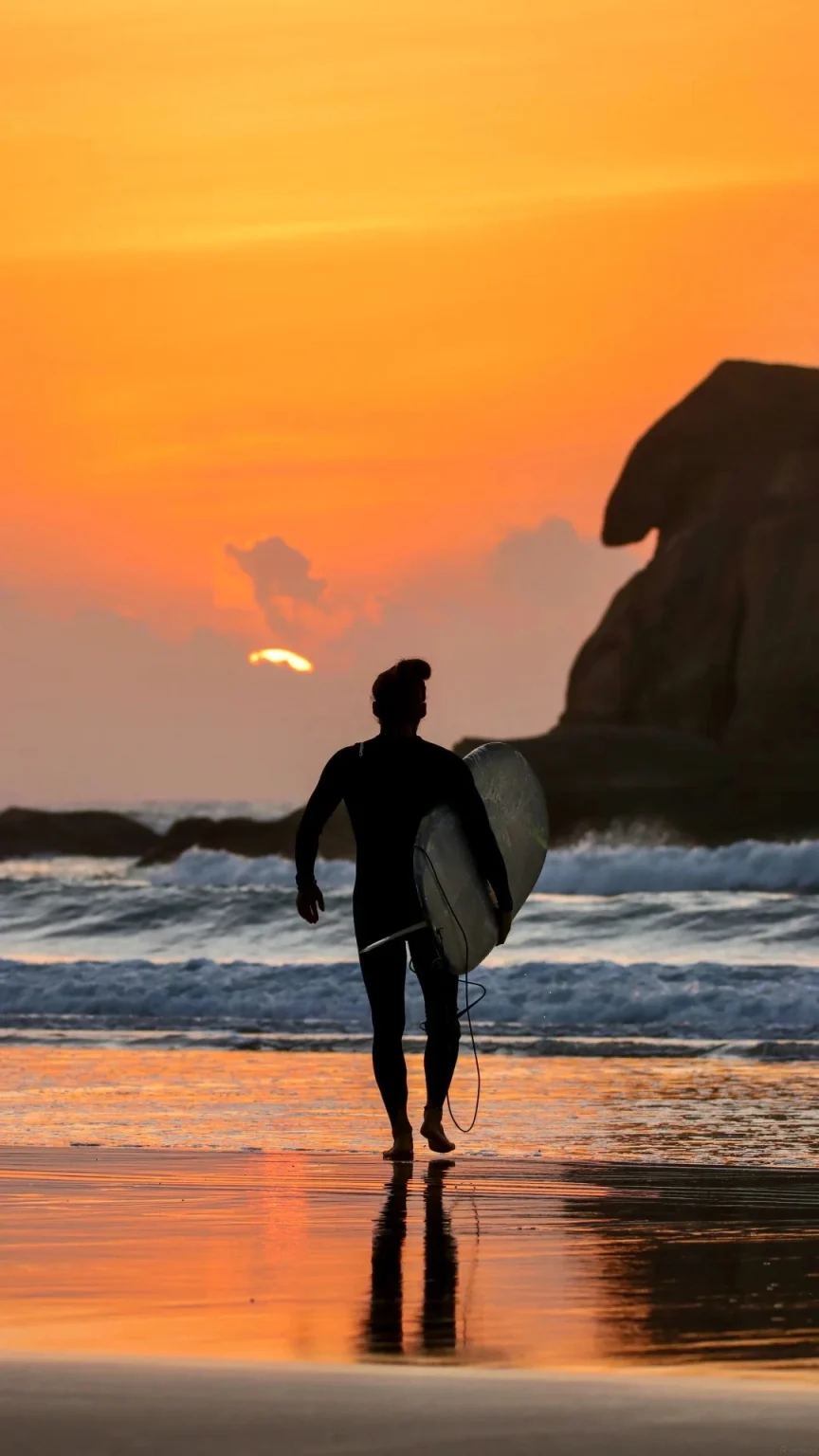 Silhouette of a surfer in a wetsuit carrying a surfboard, walking on a wet sandy beach towards the ocean. The sun is setting, casting an orange glow on the sky and reflecting on the water.
