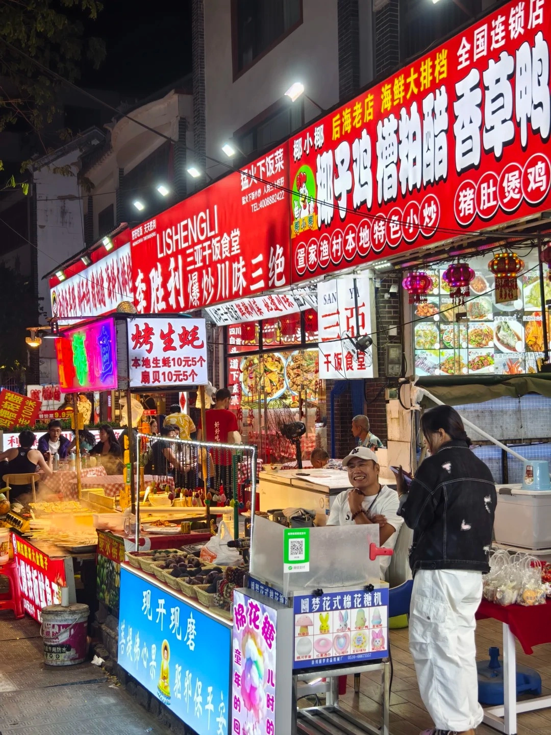 A bustling night market scene in Houhai village, with numerous brightly lit food stalls. In the foreground, a cotton candy machine is visible with a woman in white pants and a denim jacket looking at her phone. A smiling man in a white t-shirt and baseball cap sits behind the machine. Behind them, various food stalls sell skewers, fried items, and other snacks under large red and white signs with Chinese characters. People are seen eating and walking through the alleyway.