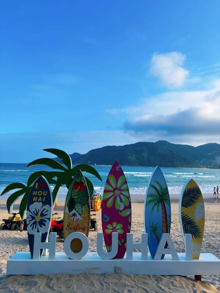 A beach scene with a large white sign spelling 'HOUHAI' in capital letters. Five decorative surfboards stand upright behind the sign. The surfboards have various colorful designs: one has a white hibiscus flower and the text 'I LOVE HOUHAI', another has tropical leaves and the text 'Go Surfing', a third has a pink background with large floral patterns, a fourth features a palm tree illustration against a blue sky, and the fifth is yellow with dark blue palm leaf patterns. Behind the surfboards and sign, there is a sandy beach, the ocean with gentle waves, and a mountainous coastline under a blue sky with scattered clouds. Several ATVs and a motorcycle are visible on the beach behind the surfboards.