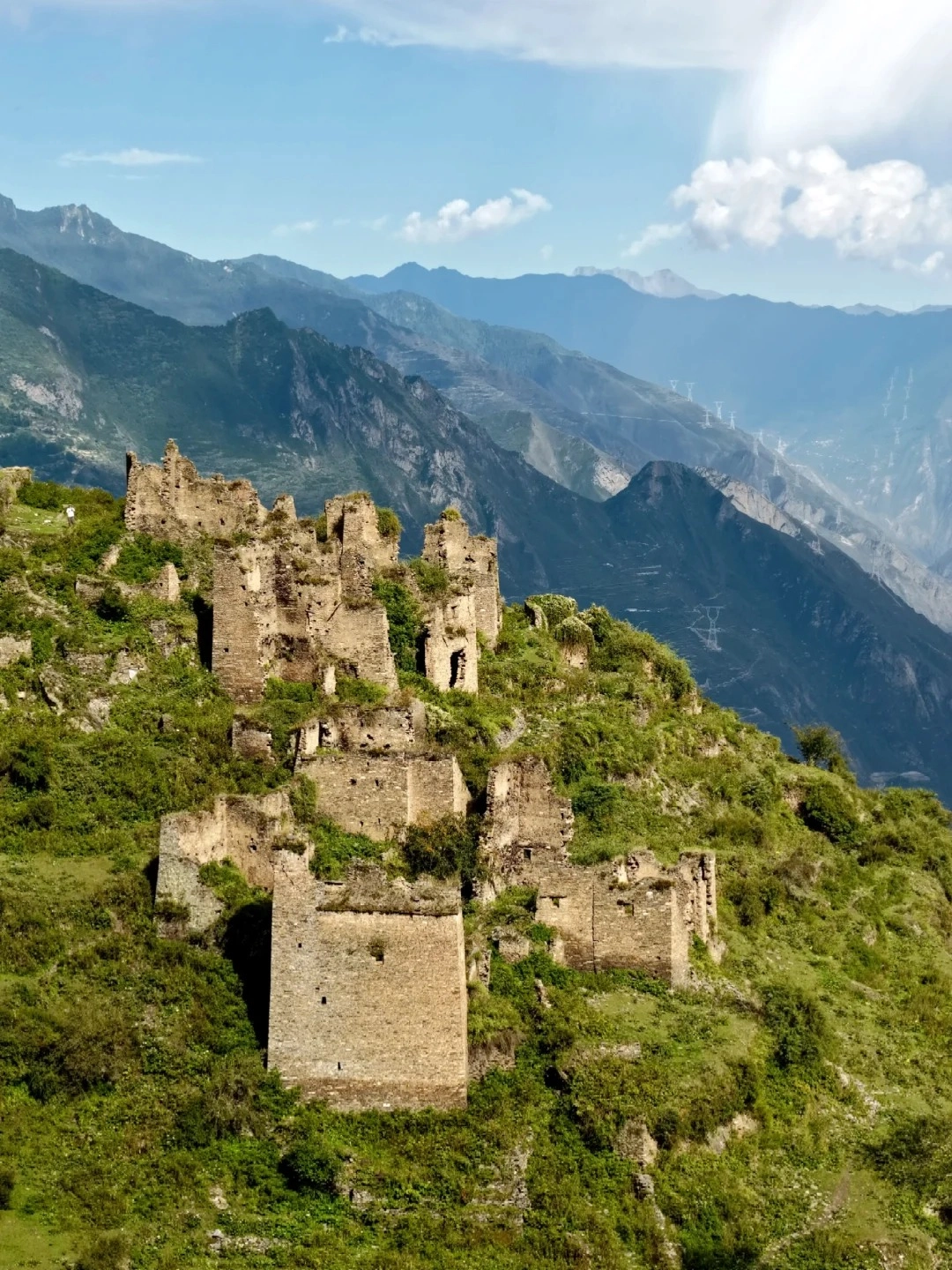 Ancient stone ruins of watchtowers on a steep, grassy mountainside, with layers of mist-shrouded mountains in the background under a blue sky with white clouds.