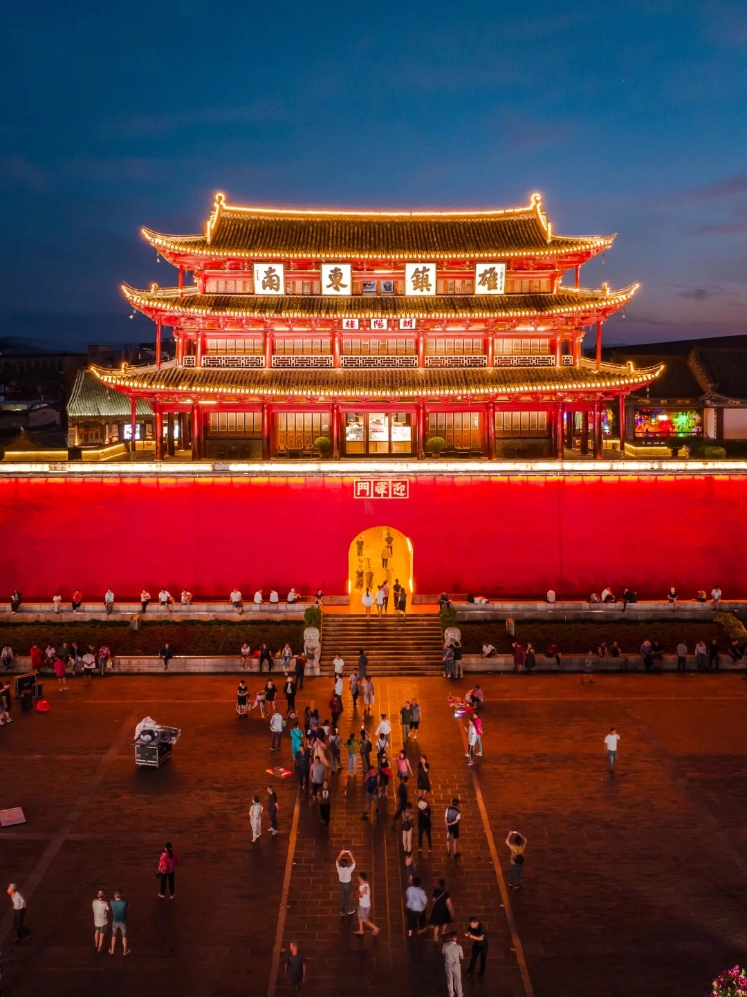 A wide-angle, elevated shot of the Chaoyang Gate tower in Jianshui Old Town at dusk. The traditional multi-tiered Chinese architectural structure is brightly illuminated with golden lights outlining its eaves and roofs. The building's facade is predominantly red, with intricate latticework on the second and third levels. A large, vibrant red wall forms the base of the structure, featuring an arched entryway leading to a set of stone stairs. Numerous people are visible walking up the stairs, entering the gate, and gathered in the large paved plaza in front of the wall. The sky above is a deep twilight blue.