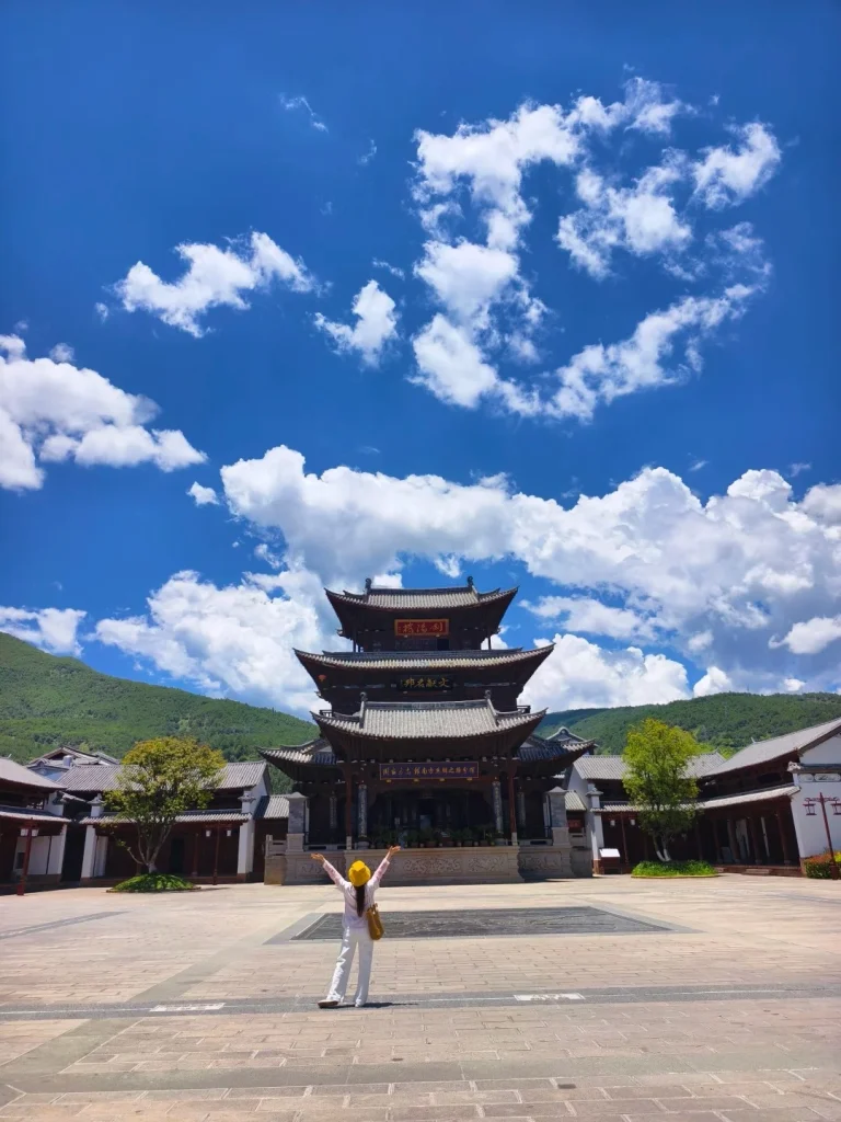 A person in a yellow hat and white clothing stands with their arms outstretched in a large, paved courtyard in front of a multi-tiered traditional Chinese temple with dark wood and gray tiled roofs. Mountains are visible in the background under a bright blue sky with scattered white clouds.