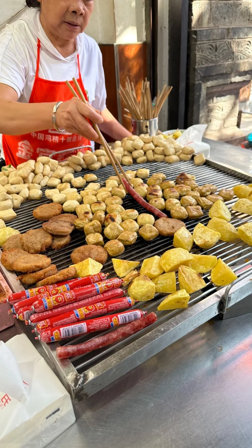 An older Asian woman wearing an orange apron uses chopsticks to turn pieces of grilled food on a metal grate. The grate holds many small, golden-brown fried tofu cubes, round meat patties, and some yellow potato chunks. Below the grill are rows of red packaged sausages and one unpacked sausage on the grill. In the background, more tofu is stacked. A metal container of wooden skewers is also visible.