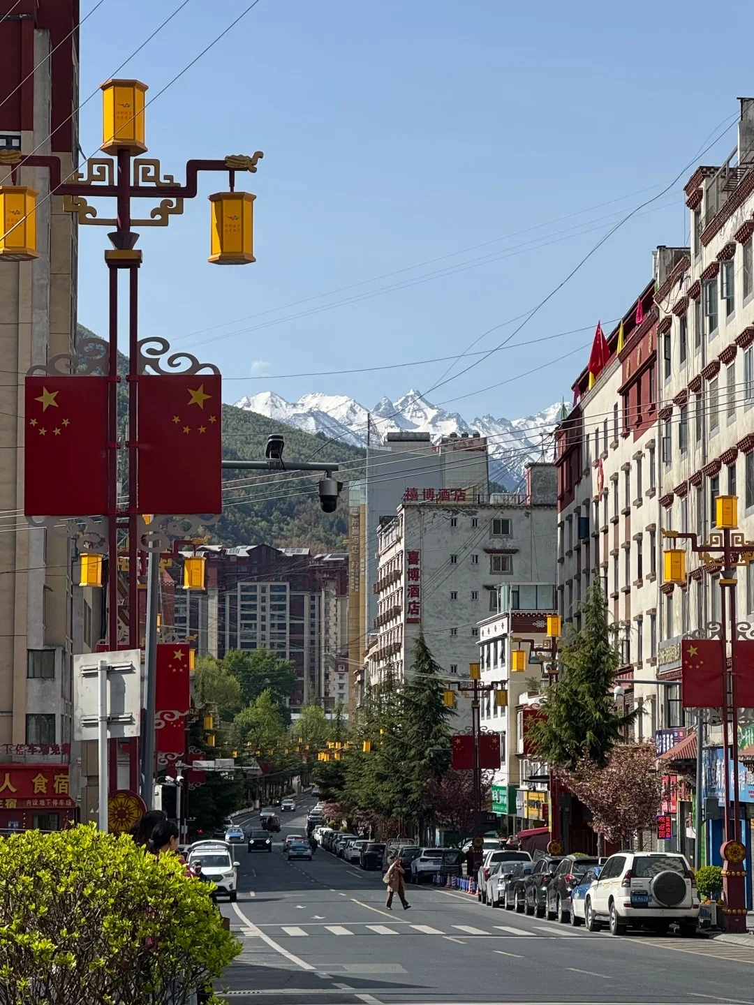 A street scene in Kangding, Sichuan, with buildings, cars, and decorative lampposts featuring Chinese flags and lanterns. Snow-capped mountains are visible in the background under a clear blue sky.
