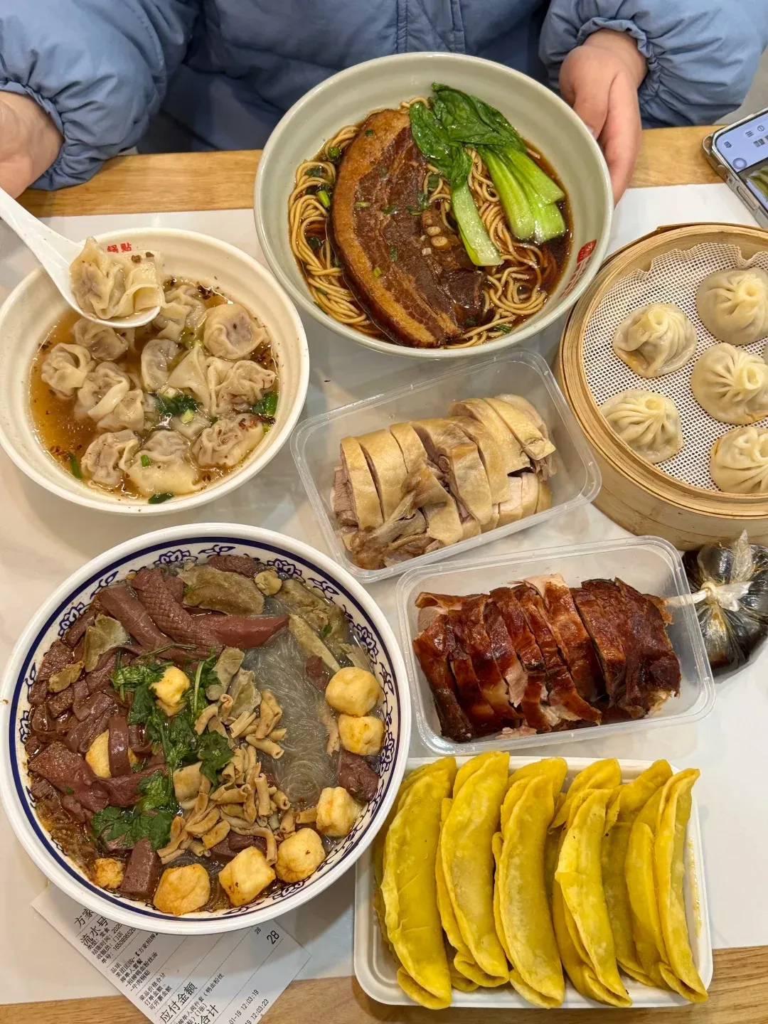 A overhead view of a breakfast spread featuring a bowl of wontons in broth, a large bowl of noodles topped with a slab of braised pork belly and greens, a bamboo steamer basket with several steamed soup dumplings (tangbao), a clear plastic container with sliced cooked chicken, a clear plastic container with sliced roasted duck, a large bowl filled with various cooked ingredients including duck blood, offal, meatballs, and noodles in broth, and a white rectangular tray holding a row of golden pan-fried dumplings (potstickers). A person's hands are visible holding a spoon lifting wontons from a bowl.