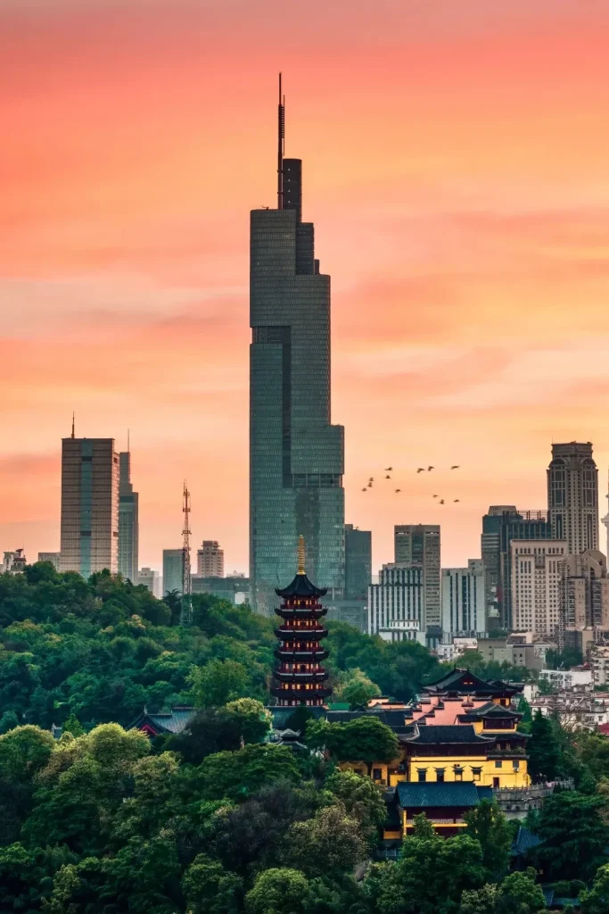 A panoramic view of the Nanjing cityscape at sunrise, with a modern skyscraper dominating the skyline and a traditional multi-tiered pagoda in the foreground, nestled among lush green trees.