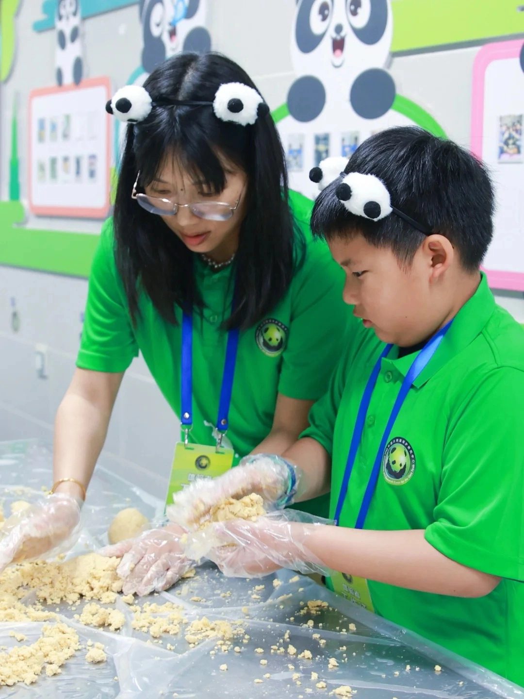 A young woman wearing glasses and a green shirt with panda ear headbands looks down at a table covered with yellow crumbly dough. Next to her, a young boy in a matching green shirt also wears panda ear headbands and is working with the dough. Both are wearing clear plastic gloves and are in the process of making something with the dough. They are in front of a wall decorated with cartoon pandas.